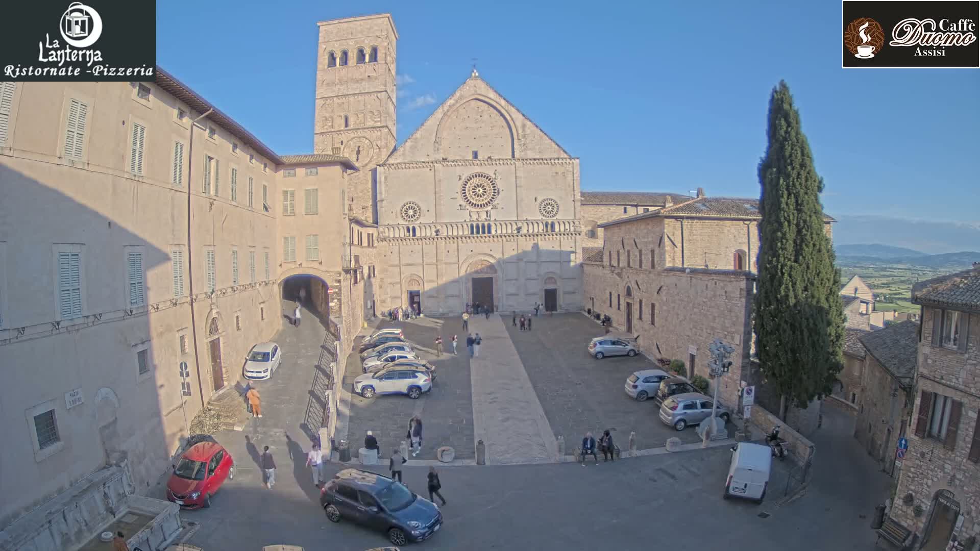 A large stone cathedral is situated in a town square, surrounded by buildings, parked cars, and a few people; the weather is sunny and clear.