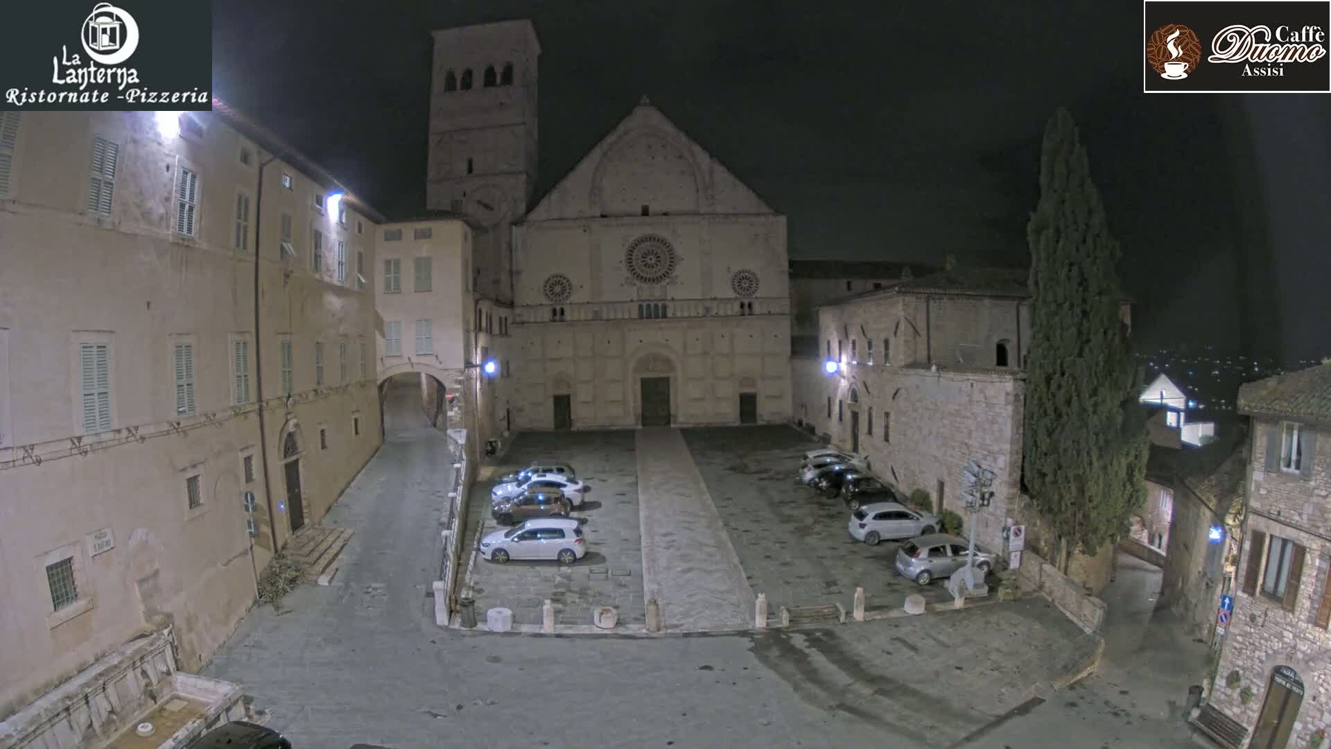 A nighttime, outdoor view of a town square with several cars parked in front of a large stone building, surrounded by other buildings.