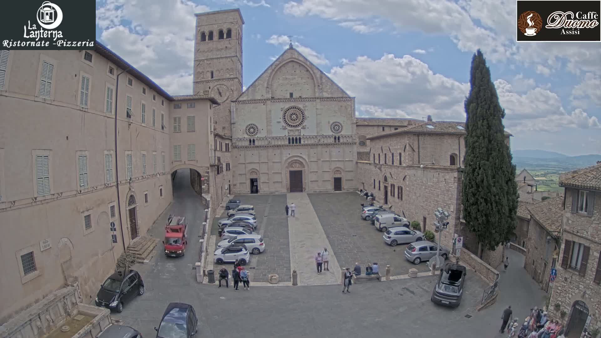 A large stone cathedral is situated in a town square with cars parked, pedestrians walking, and other buildings surrounding it under a partly cloudy sky.
