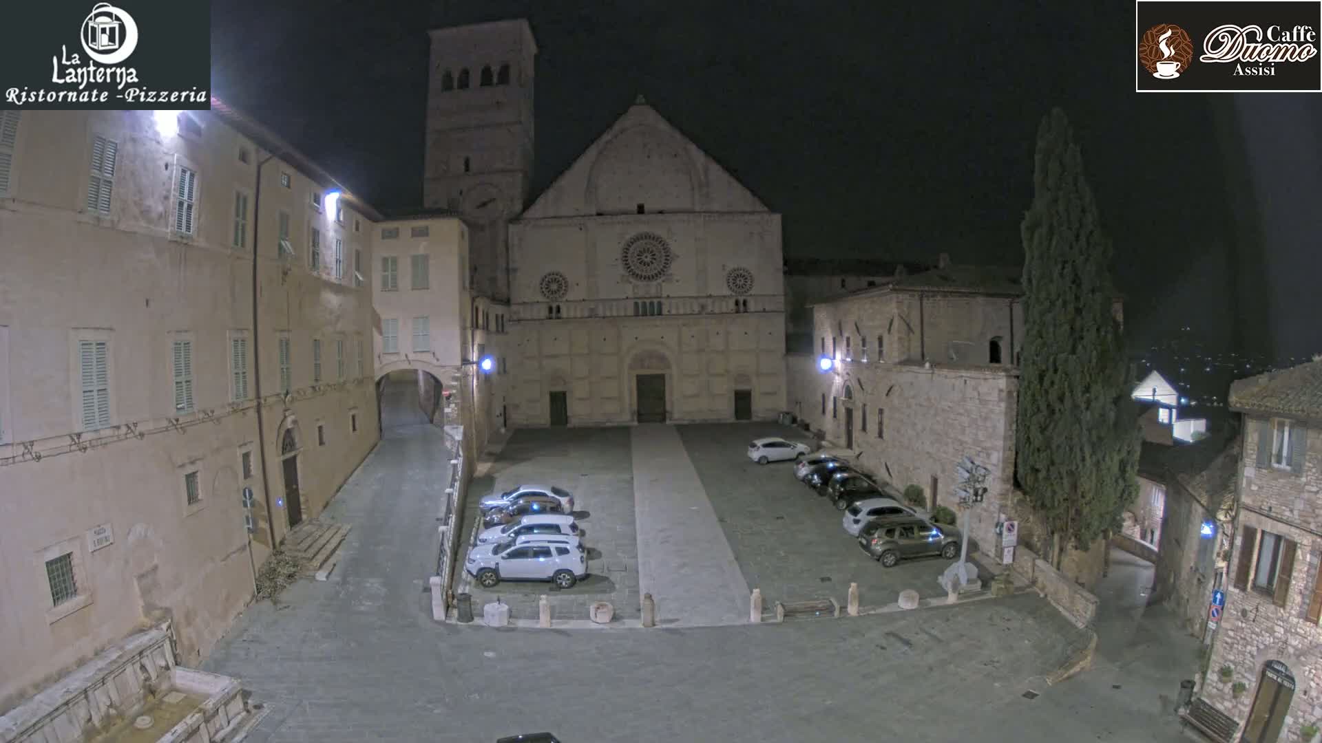 A nighttime view of a town square with several cars parked in front of a large stone building, under a clear, dark sky.