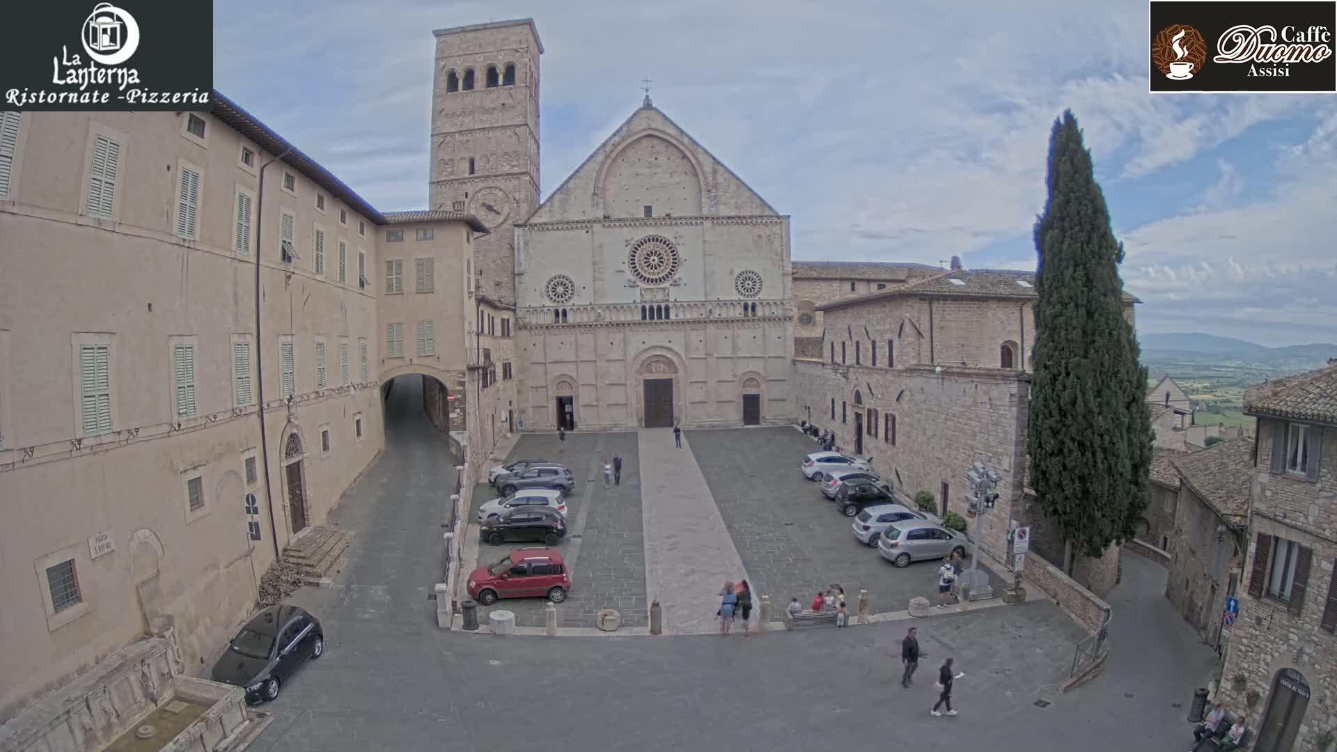 A large stone cathedral sits in a town square with several cars parked, surrounded by buildings under a partly cloudy sky.