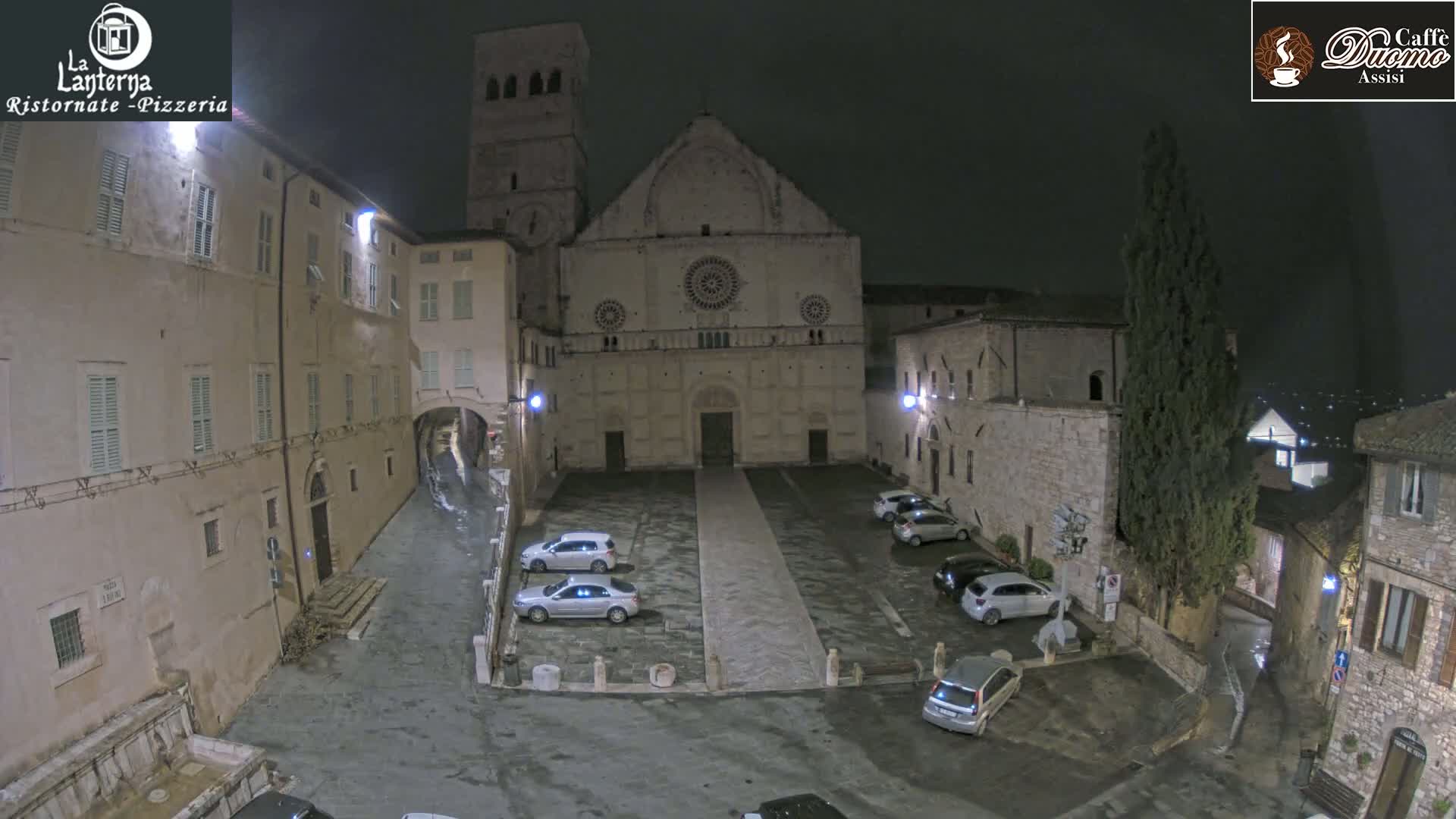 A nighttime view of a town square with a large stone church, several parked cars, and wet cobblestone streets.