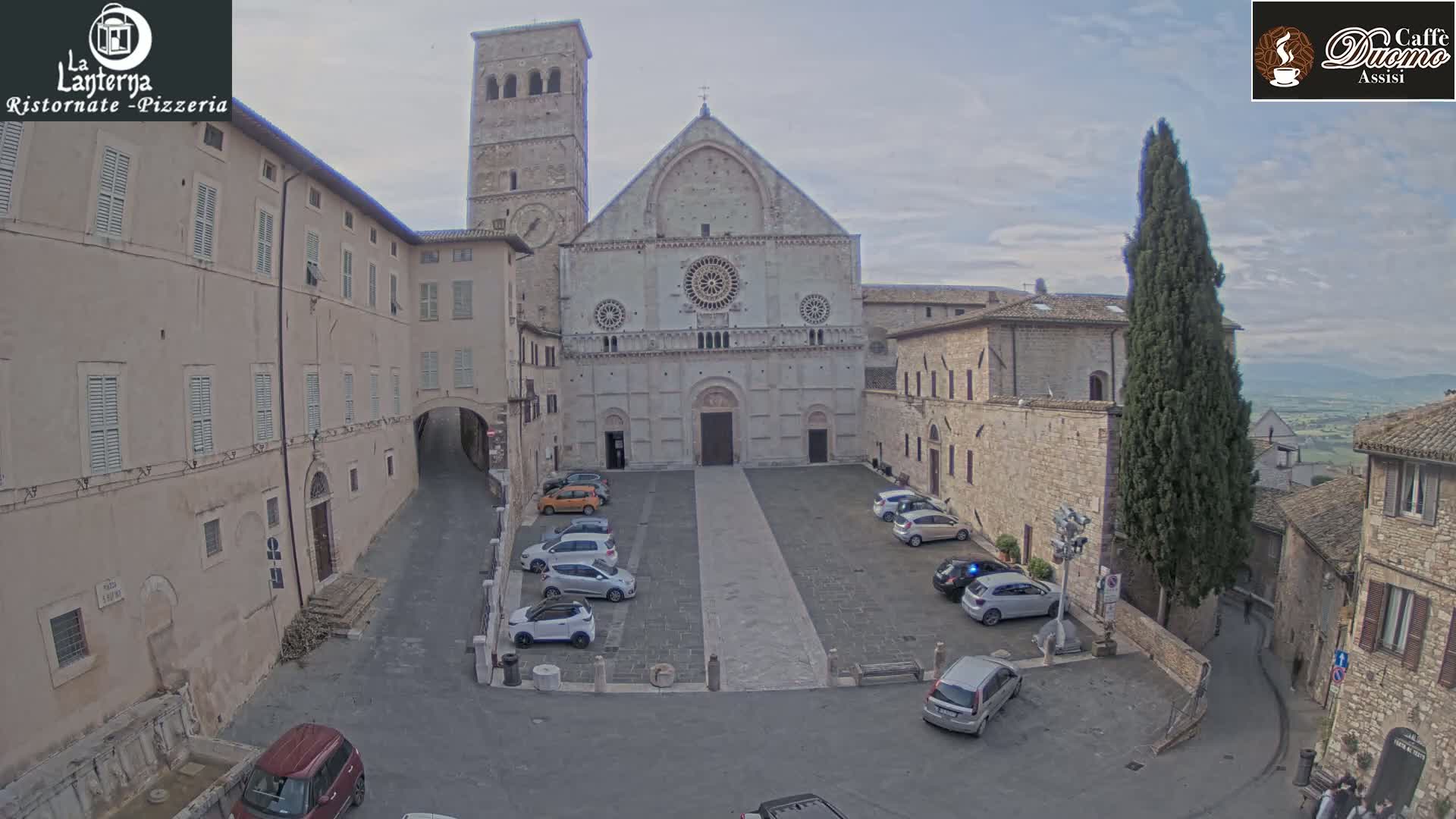 A large, light-colored stone church with a bell tower stands in a town square, where several cars are parked under an overcast sky.