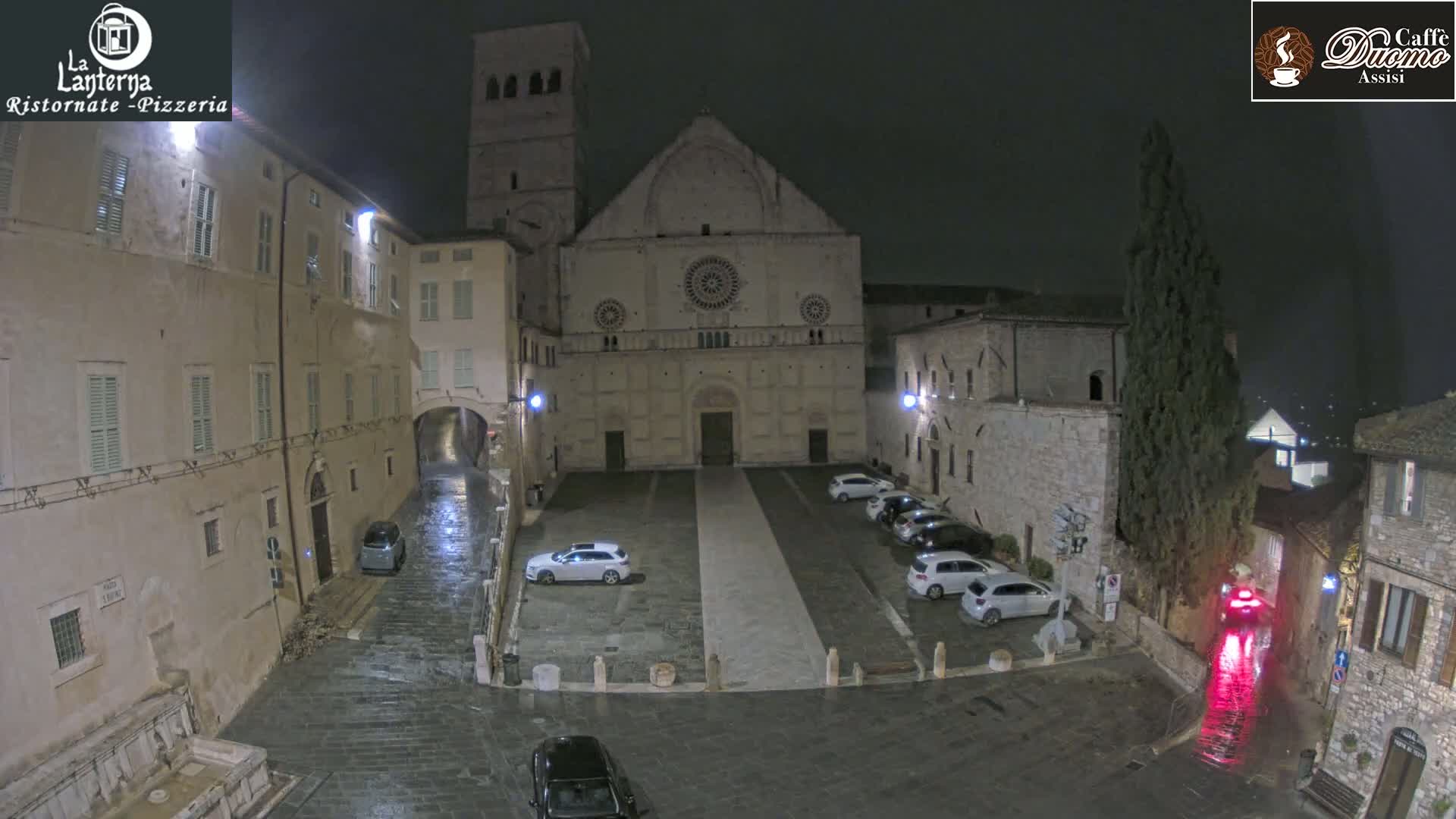 A nighttime, wet, town square with several cars parked in front of a large stone building, surrounded by other buildings.