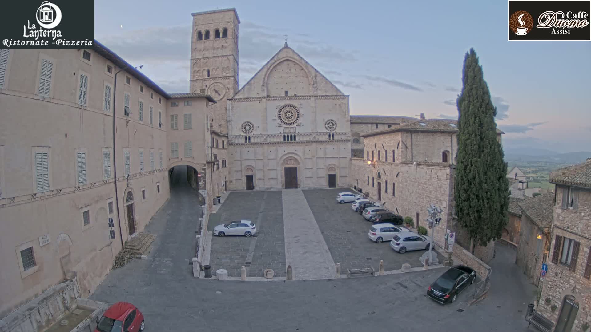 A large stone cathedral sits in a town square with several cars parked, under a twilight sky.