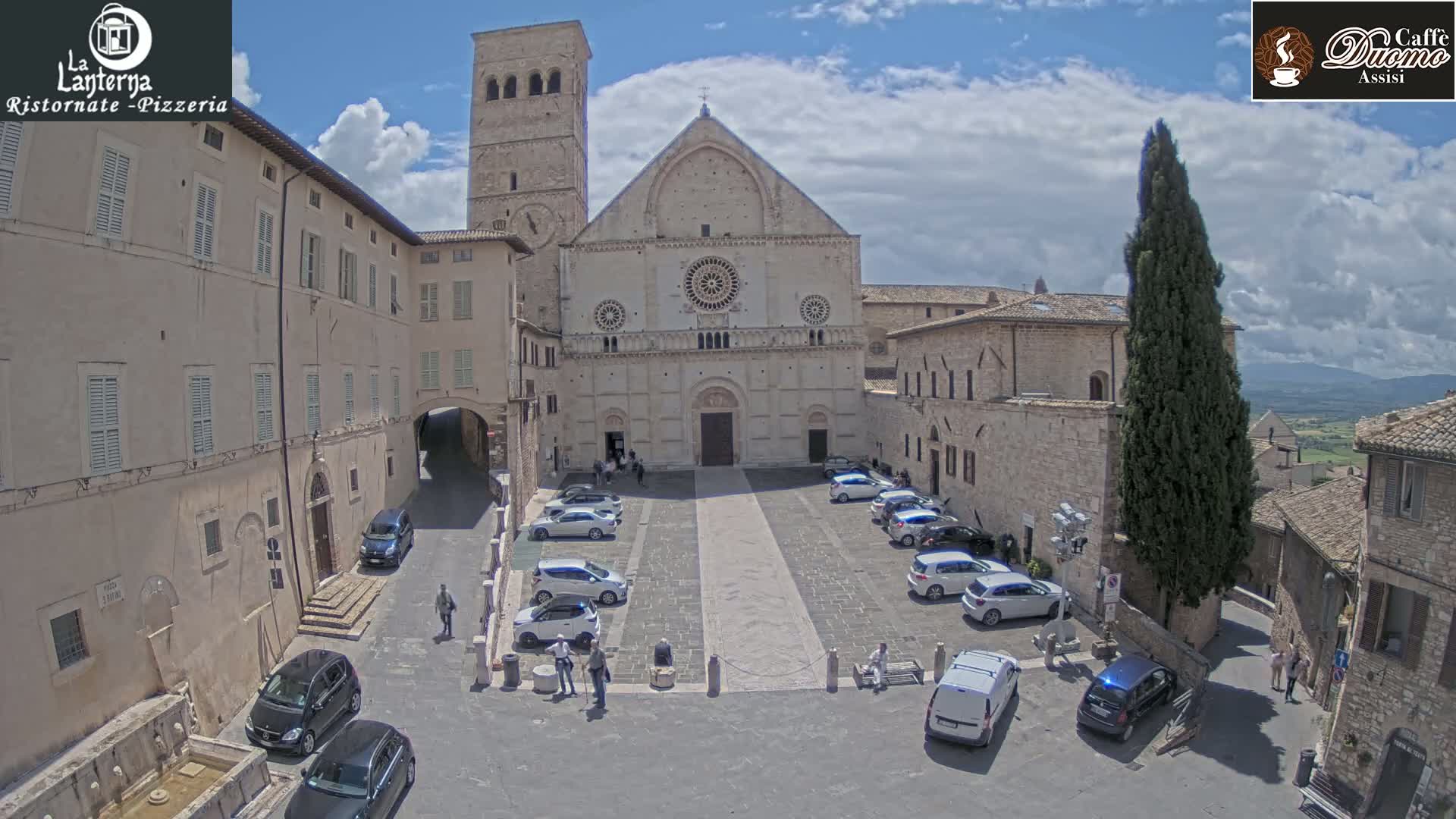 A large stone church sits in a town square on a partly cloudy day, surrounded by buildings and parked cars.