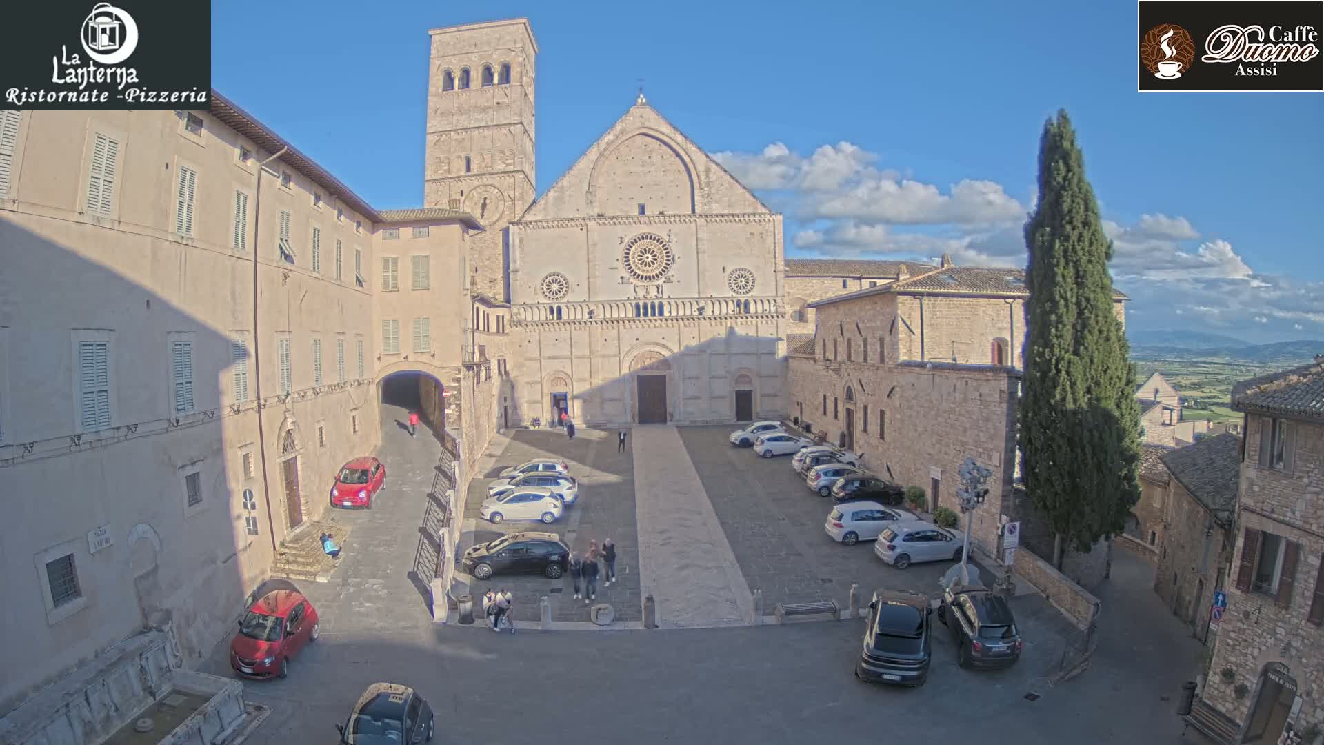 A large stone church stands in a sunny plaza surrounded by old buildings, with several cars parked and a few people walking.