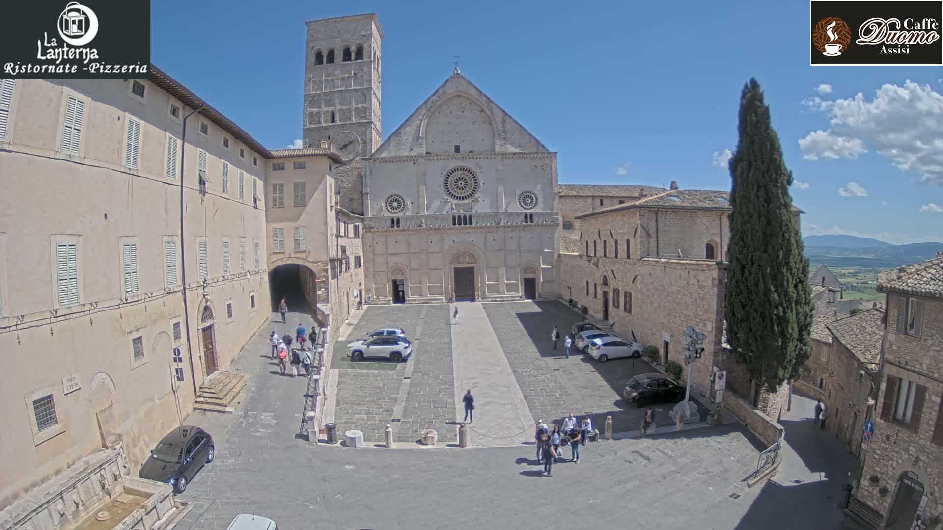 A large stone church sits in a town square on a sunny day, surrounded by buildings and a few cars and people.