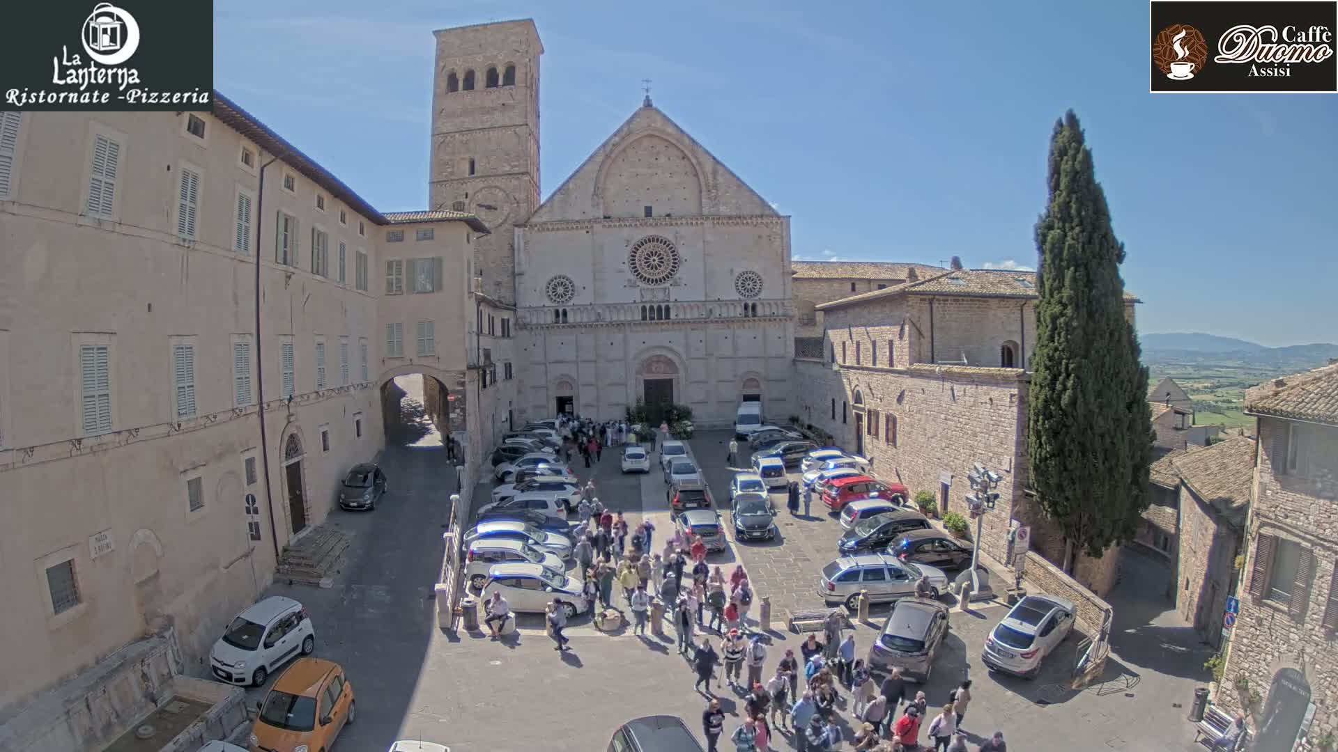 A large stone cathedral stands in a town square filled with parked cars and a crowd of people on a sunny day.