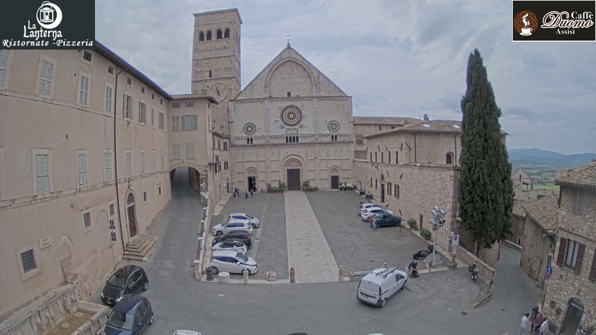 A large stone church sits in a town square with several cars parked, under an overcast sky.