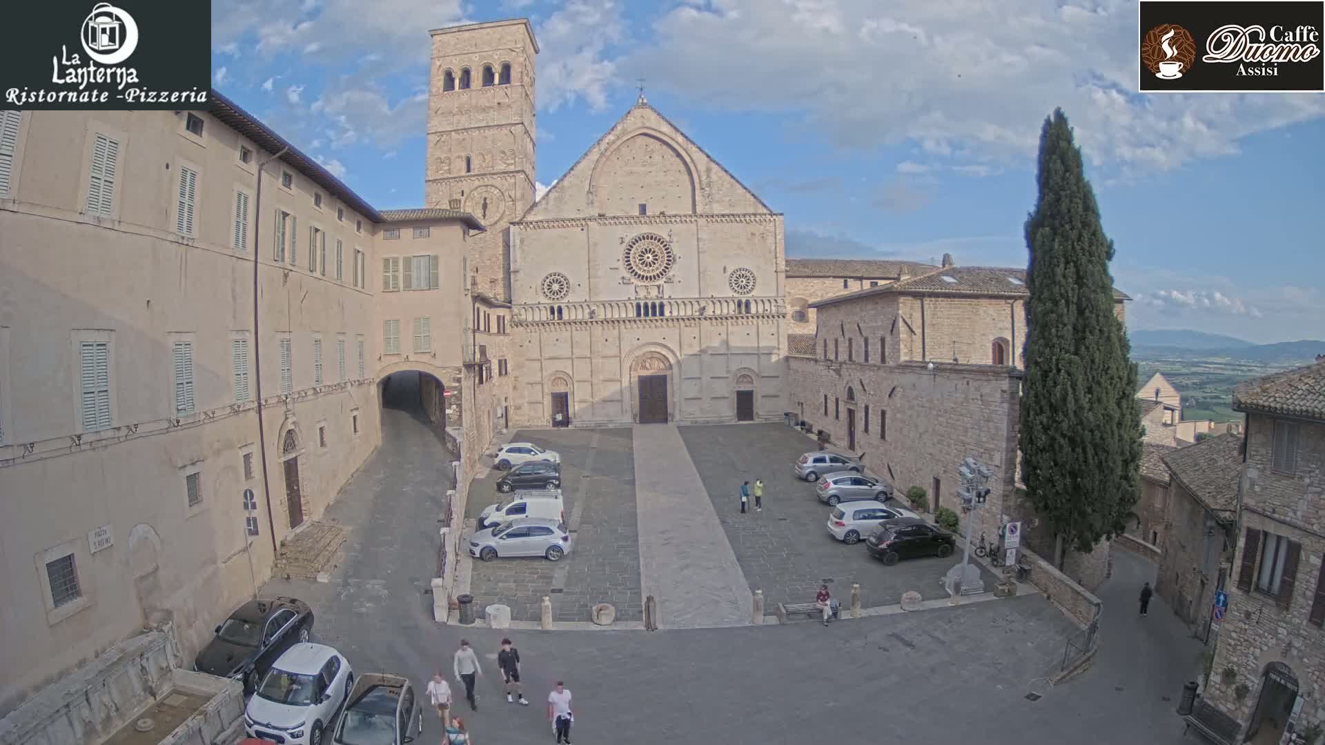 A cathedral stands in a town square with several cars parked, people walking around, and a tall, slender tree nearby under a partly cloudy sky.