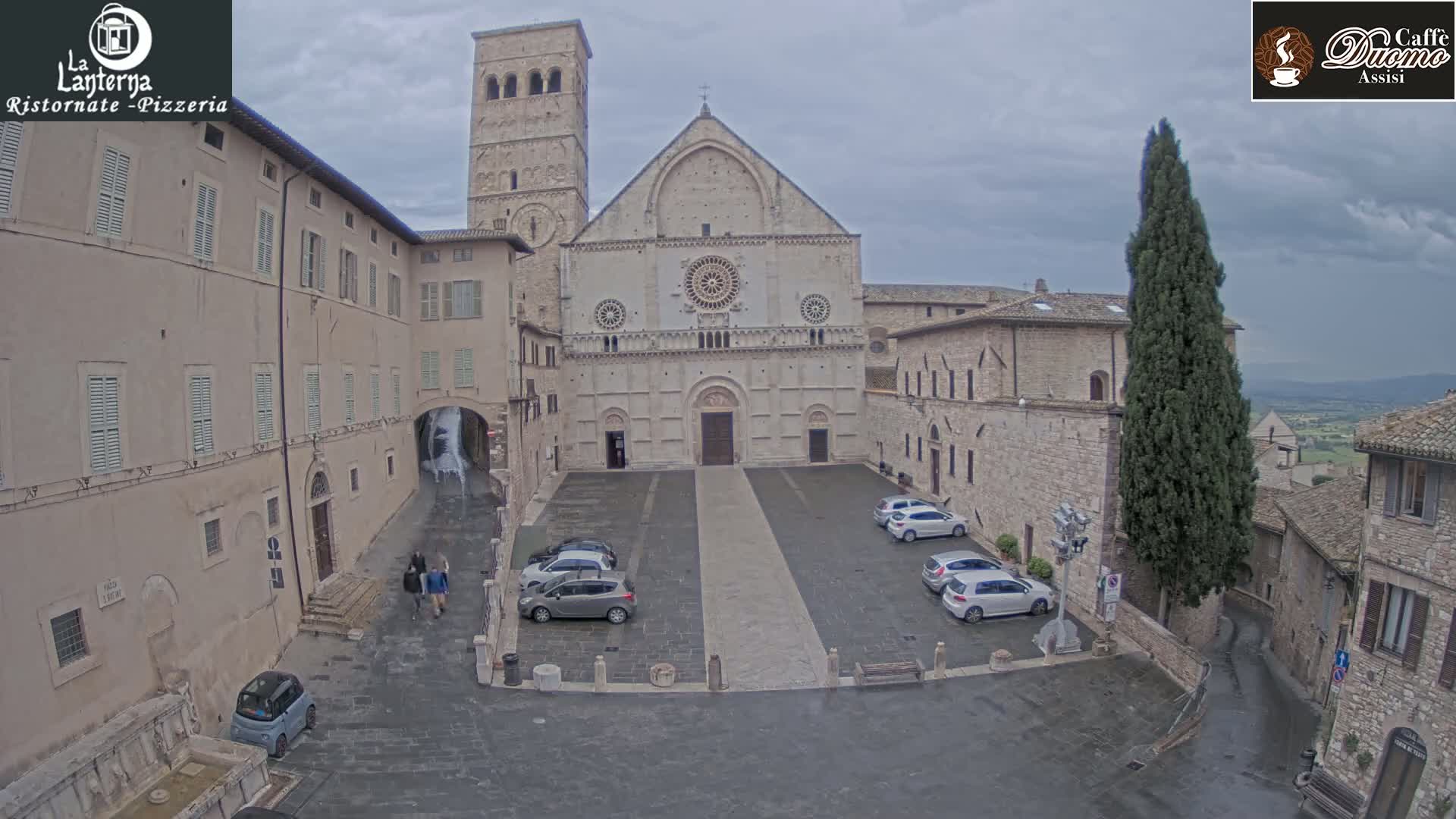 A large stone church is situated in a town square with several cars parked, under an overcast sky.