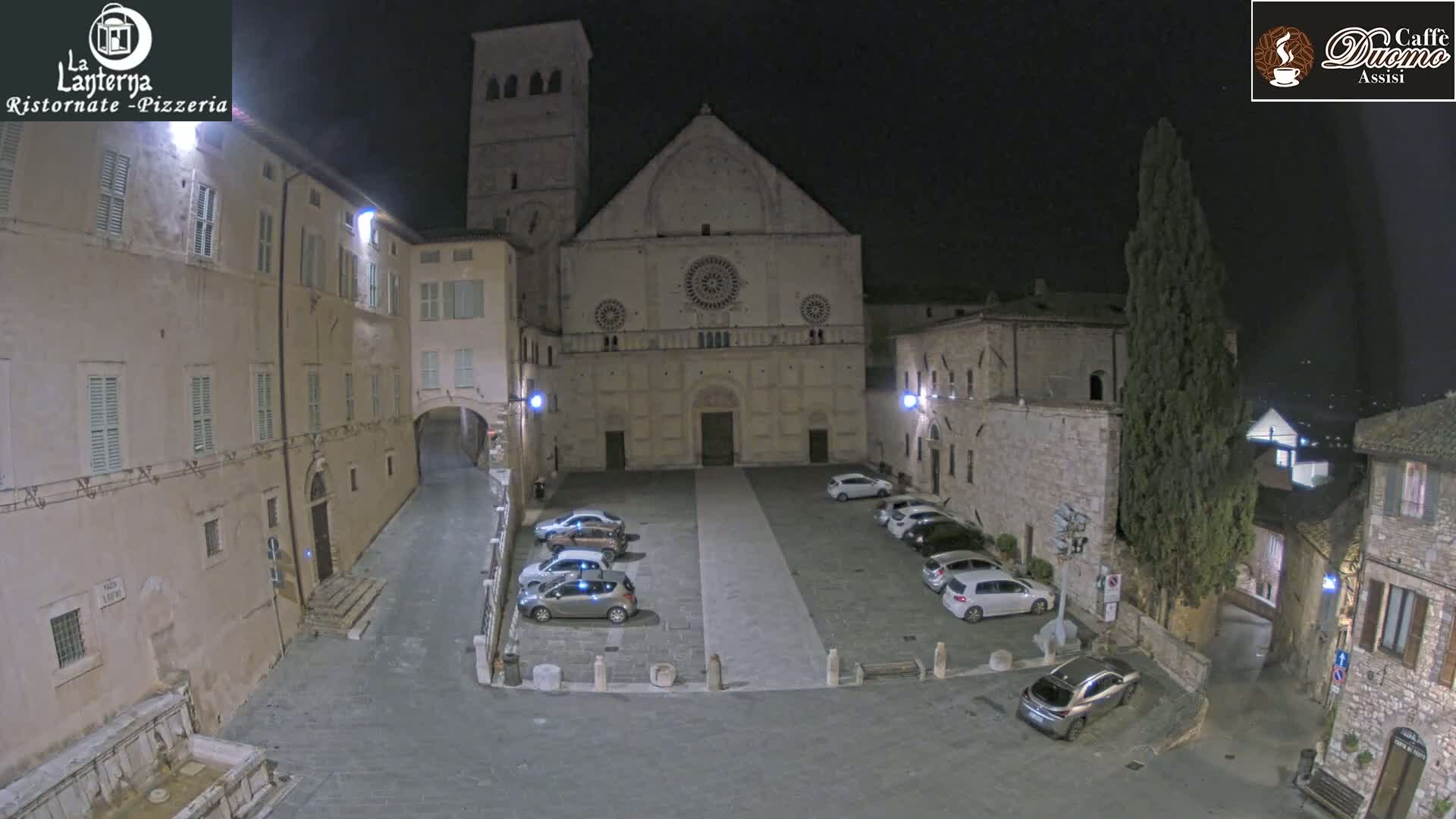 A nighttime view of a town square with parked cars in front of a large stone church, under a clear, dark sky.