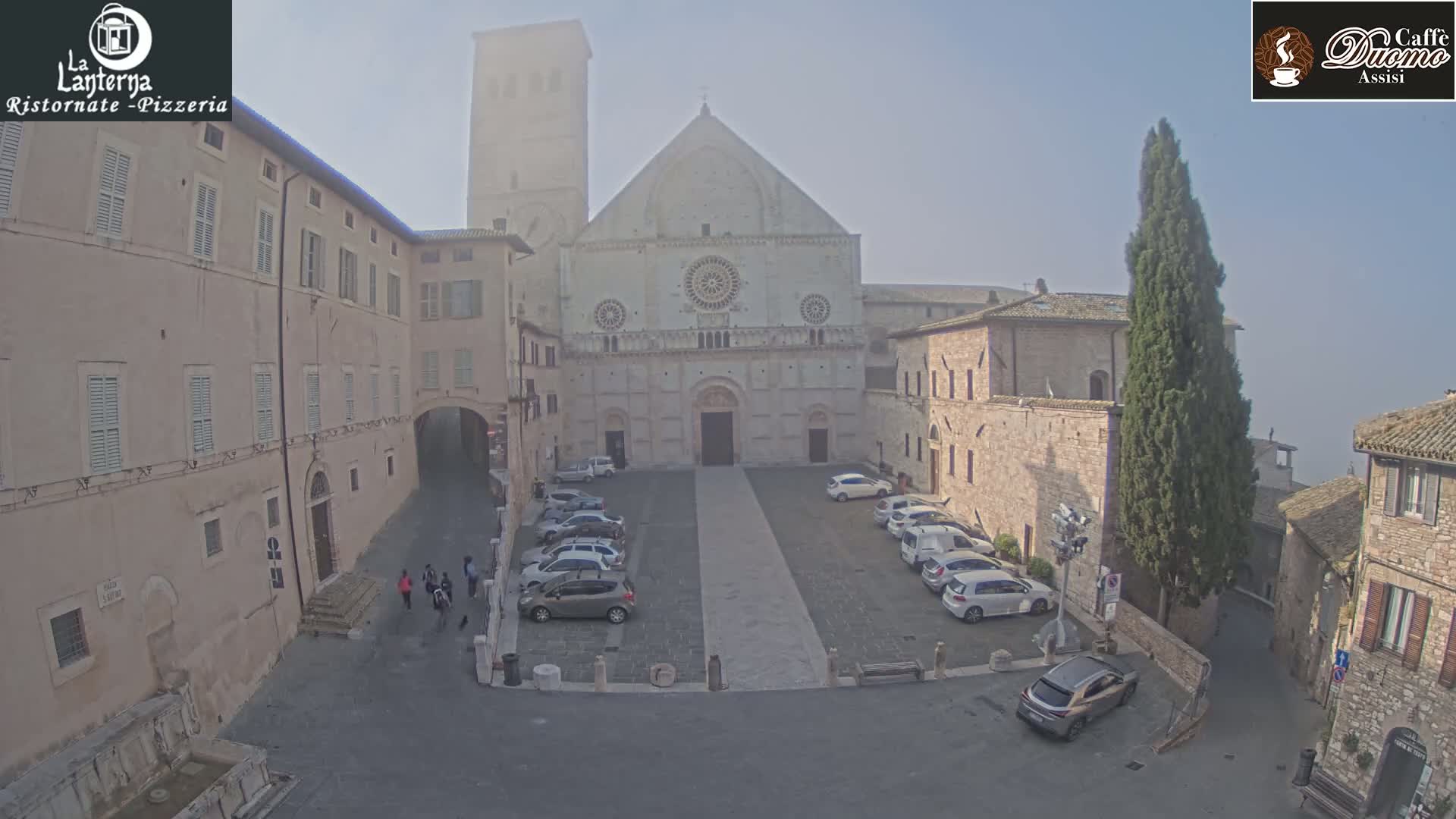 A large, light-colored cathedral faces a square with several parked cars; the scene is slightly hazy under a mostly clear sky.