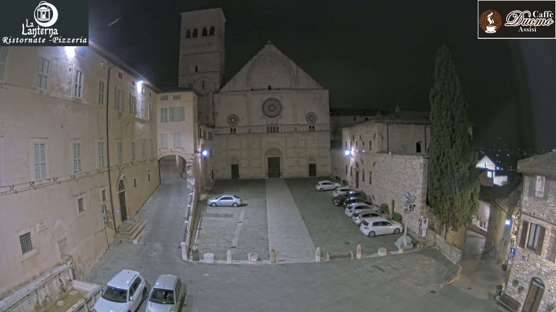 A nighttime view of a town square, featuring a large stone church at the far end, several parked cars, and surrounding buildings under a clear sky.