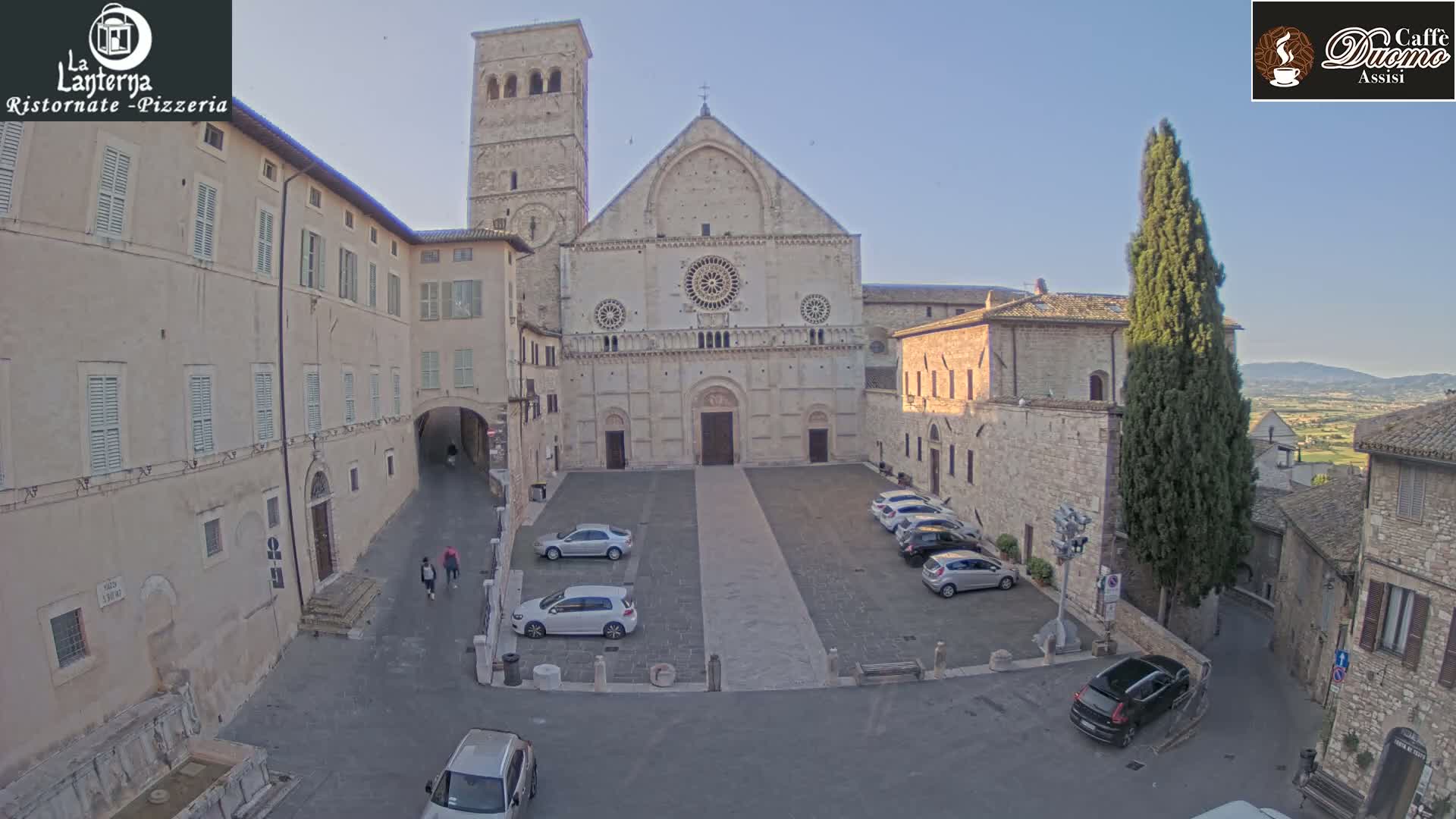 A large stone church and surrounding buildings are shown in a town square on a sunny day, with several cars parked in the square.