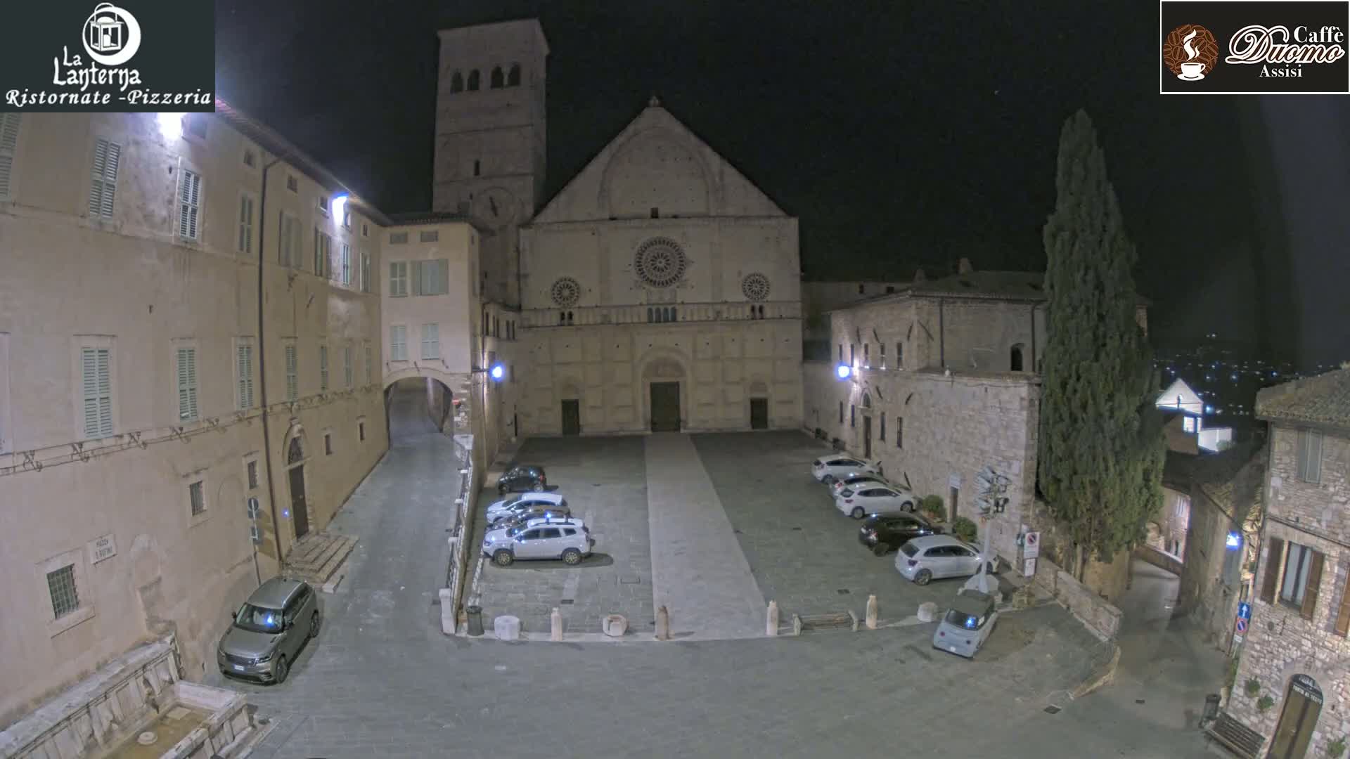 A nighttime view of a town square with parked cars in front of a large stone cathedral, under a clear, dark sky.
