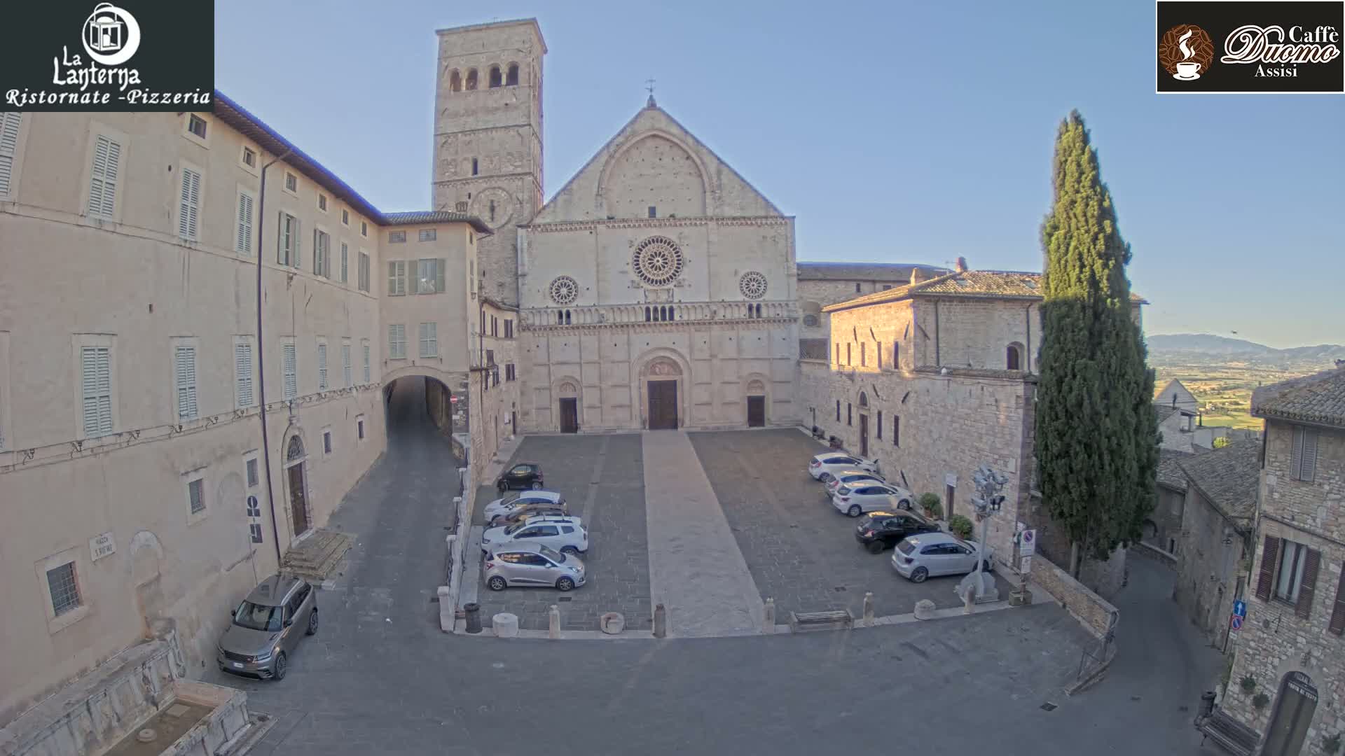 A large stone cathedral with a tall bell tower stands in a town square with several parked cars under a clear, sunny sky.