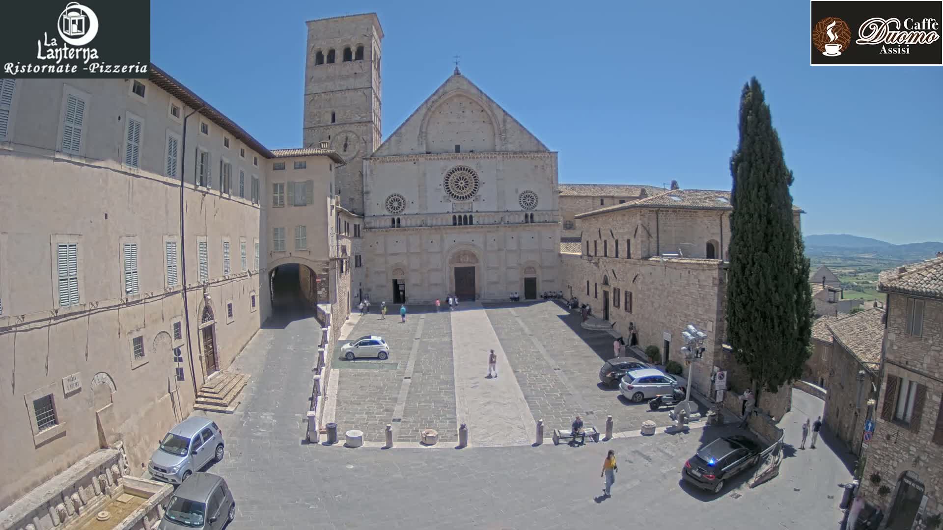 A large stone cathedral sits in a town square on a sunny day, surrounded by old stone buildings and several cars parked nearby.