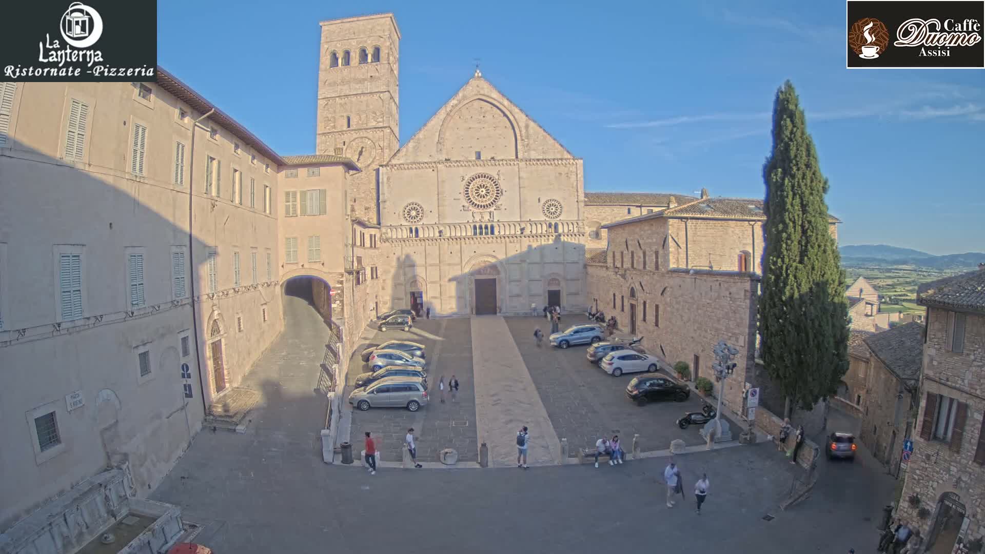 A large stone church with a plaza in front, surrounded by buildings, under a clear blue sky.