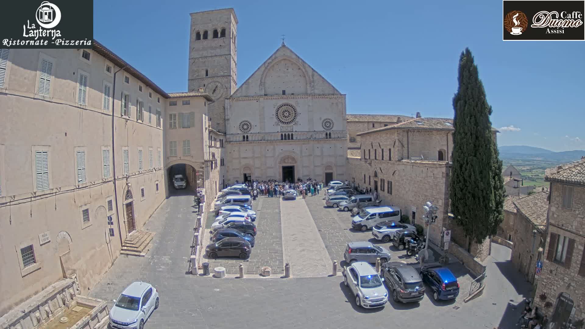 A large stone church is surrounded by a town square with parked cars and a crowd of people gathered in front of it under a clear, sunny sky.
