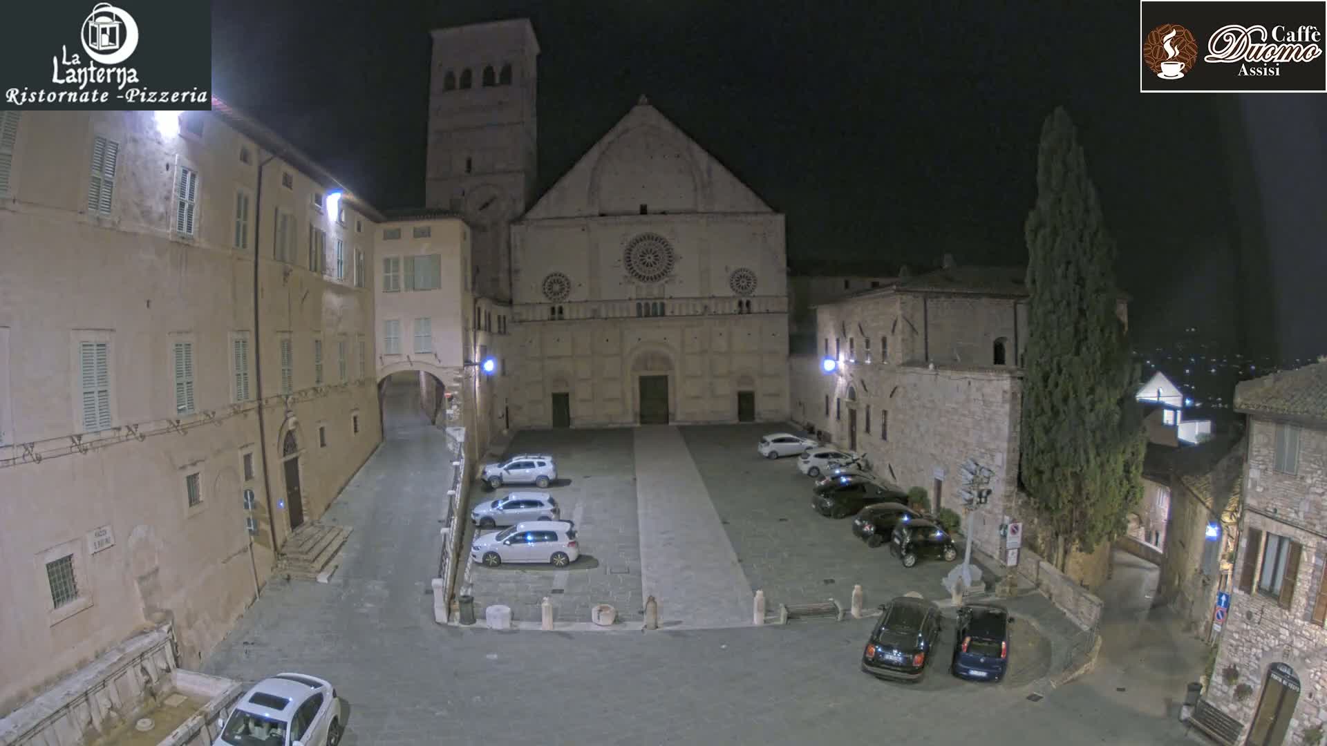 A nighttime view of a town square with several cars parked in front of a large stone church, under a clear, dark sky.
