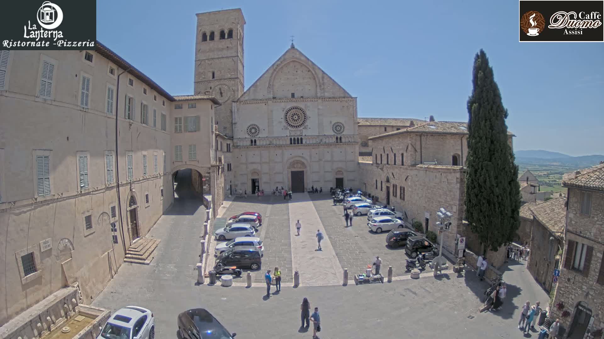 A large stone church is situated in a town square with cars parked and people walking around on a sunny day.