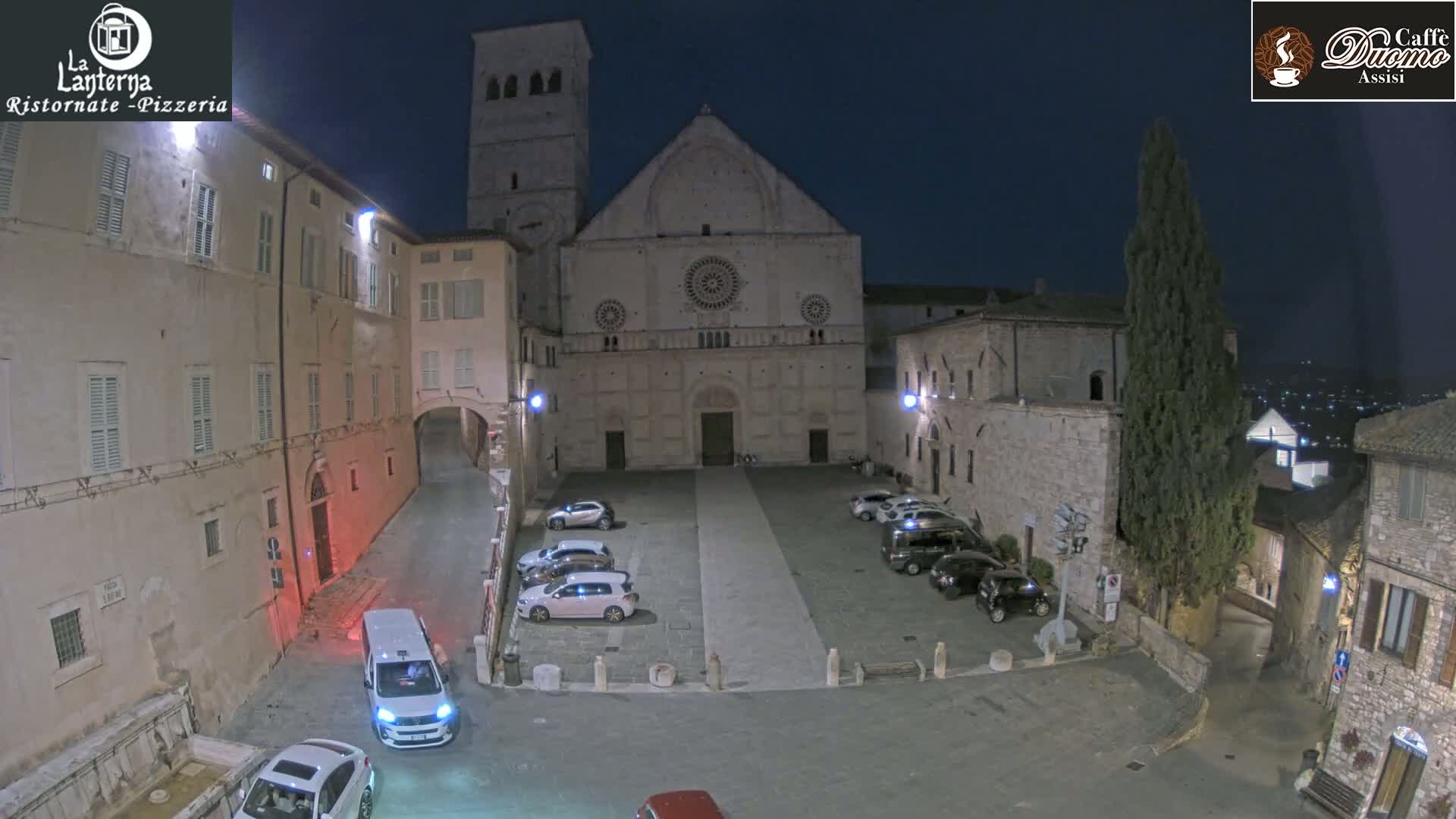 A nighttime view of a town square with several cars parked, centered on a large stone church, surrounded by old stone buildings under a dark sky.