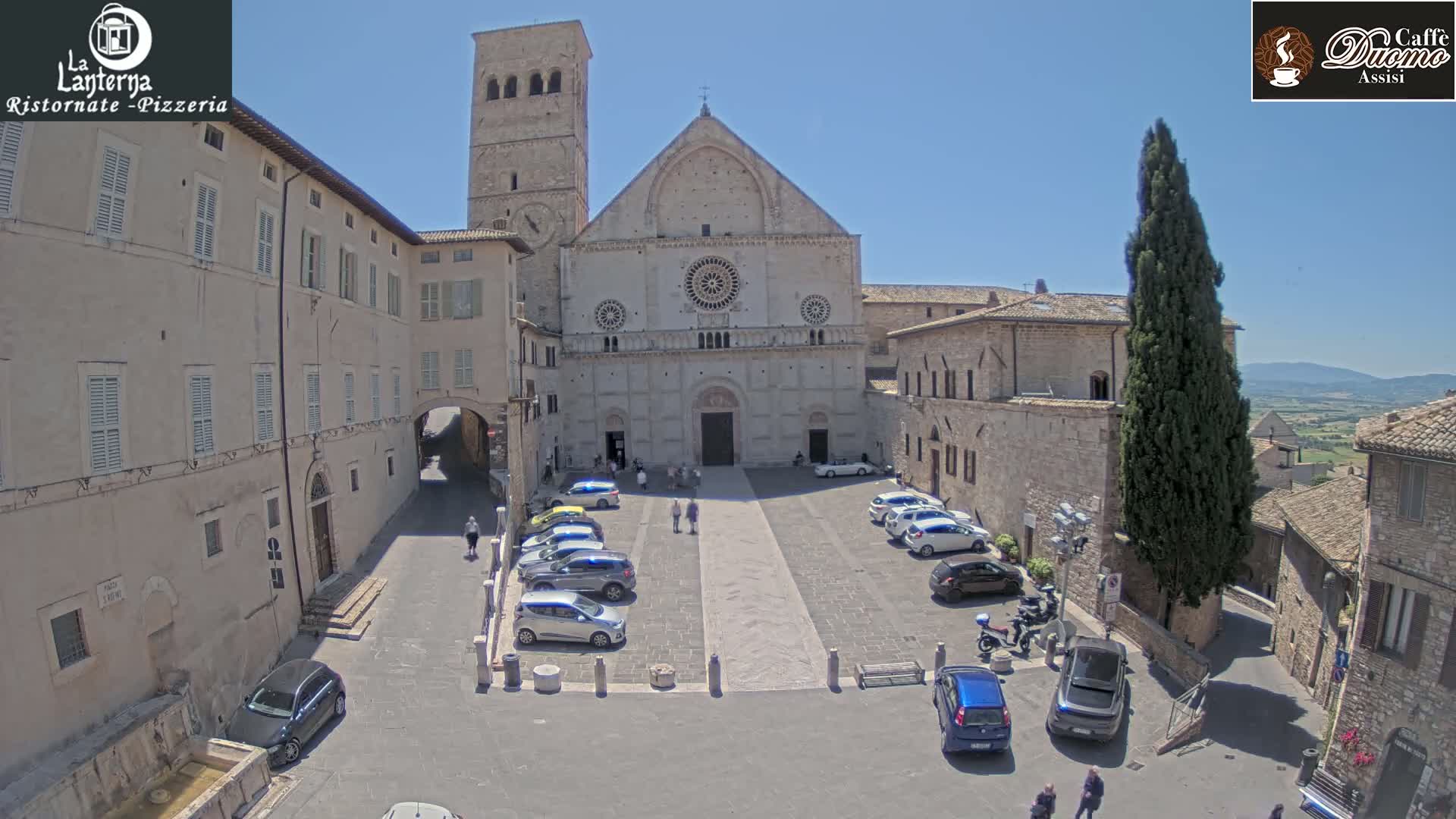 A large stone cathedral sits in a town square with cars parked around it, under a clear blue sky.