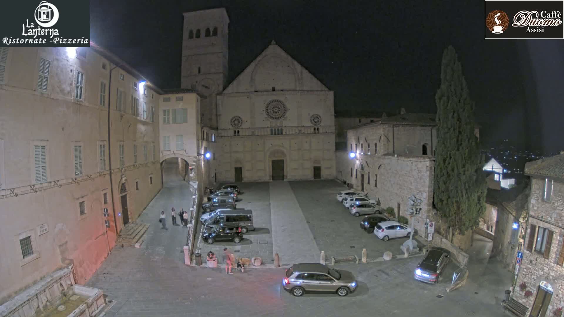 A nighttime aerial view of a town square with parked cars in front of a large stone cathedral under a clear, dark sky.