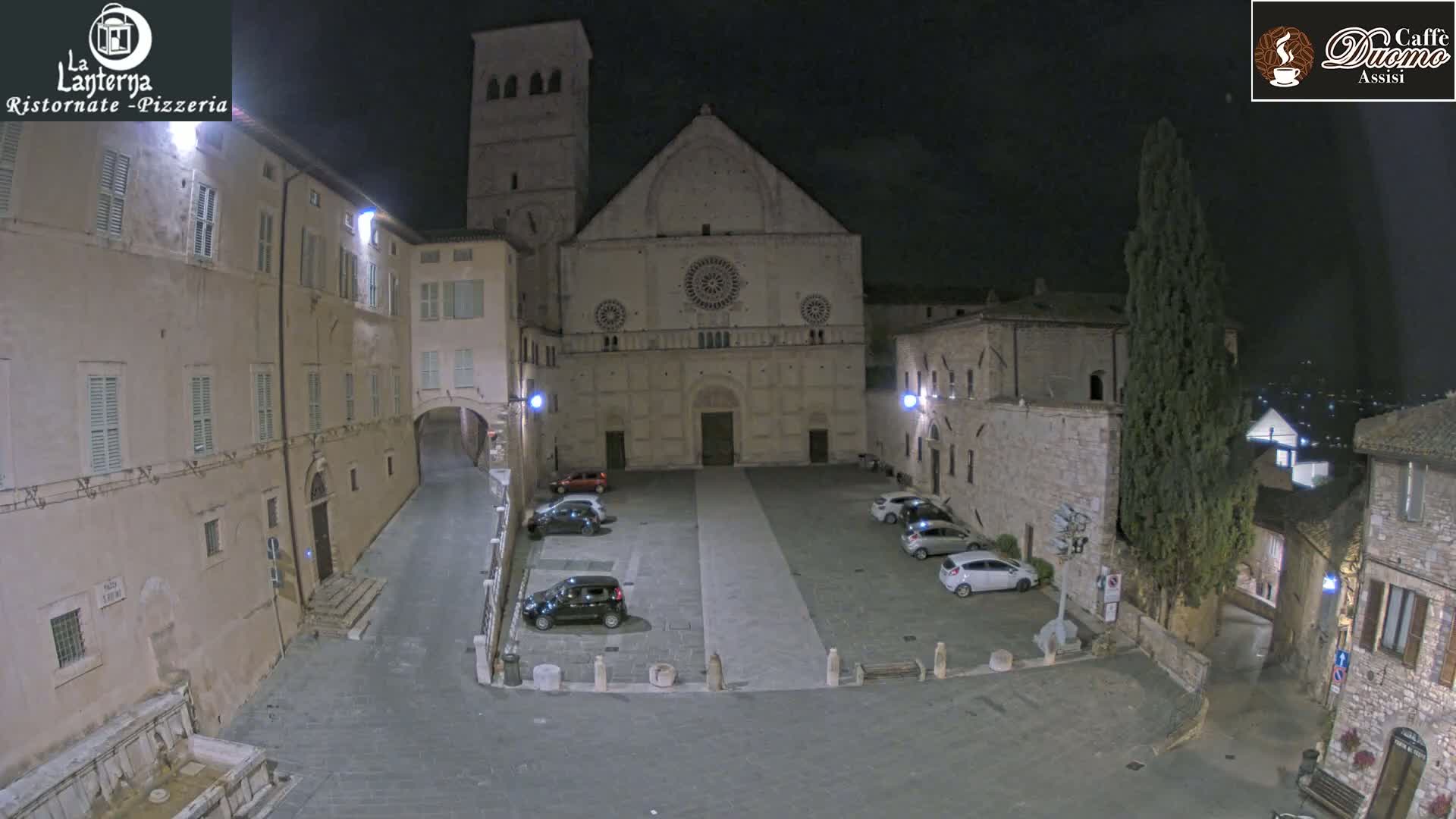 A nighttime view of a town square with a large stone church, several parked cars, and buildings illuminated by streetlights.