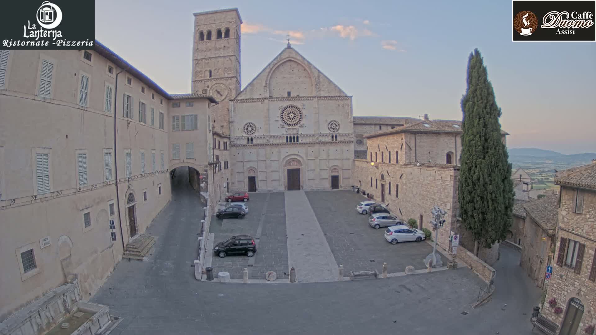 A large stone cathedral sits in a town square with several cars parked and surrounded by other stone buildings under a mostly clear evening sky.