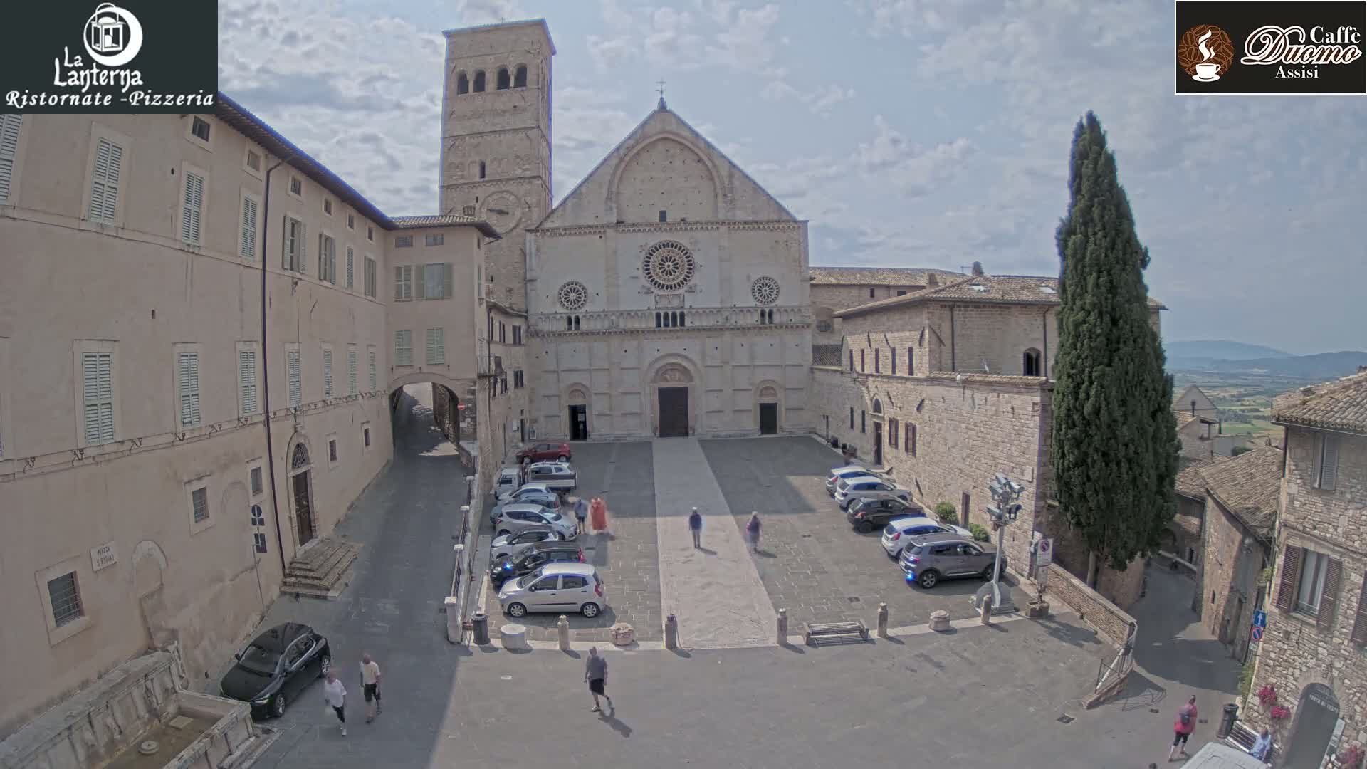 A large stone cathedral stands in a town square, surrounded by buildings and parked cars under a partly cloudy sky.
