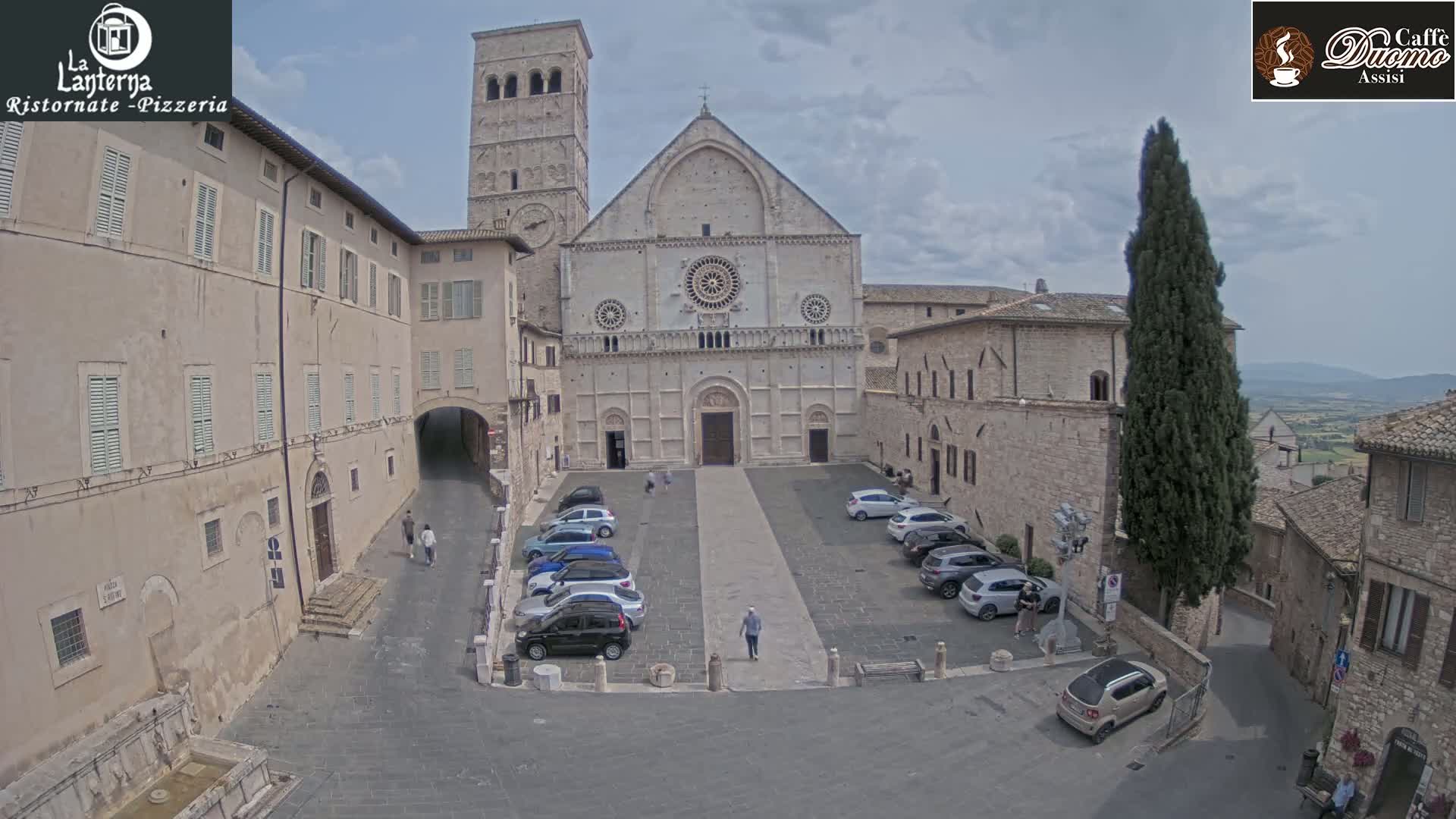 A large stone cathedral sits in a town square with cars parked around its perimeter on a partly cloudy day.