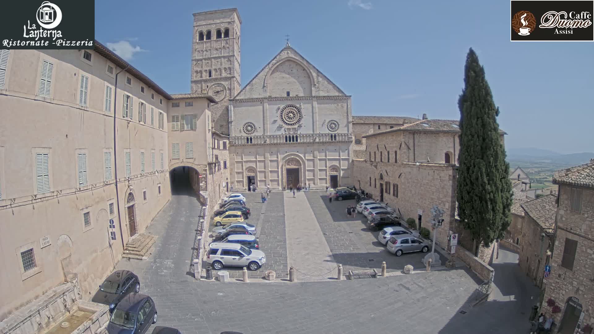 A large stone cathedral is situated in a town square, surrounded by older stone buildings, under a clear, sunny sky; cars are parked in the square.