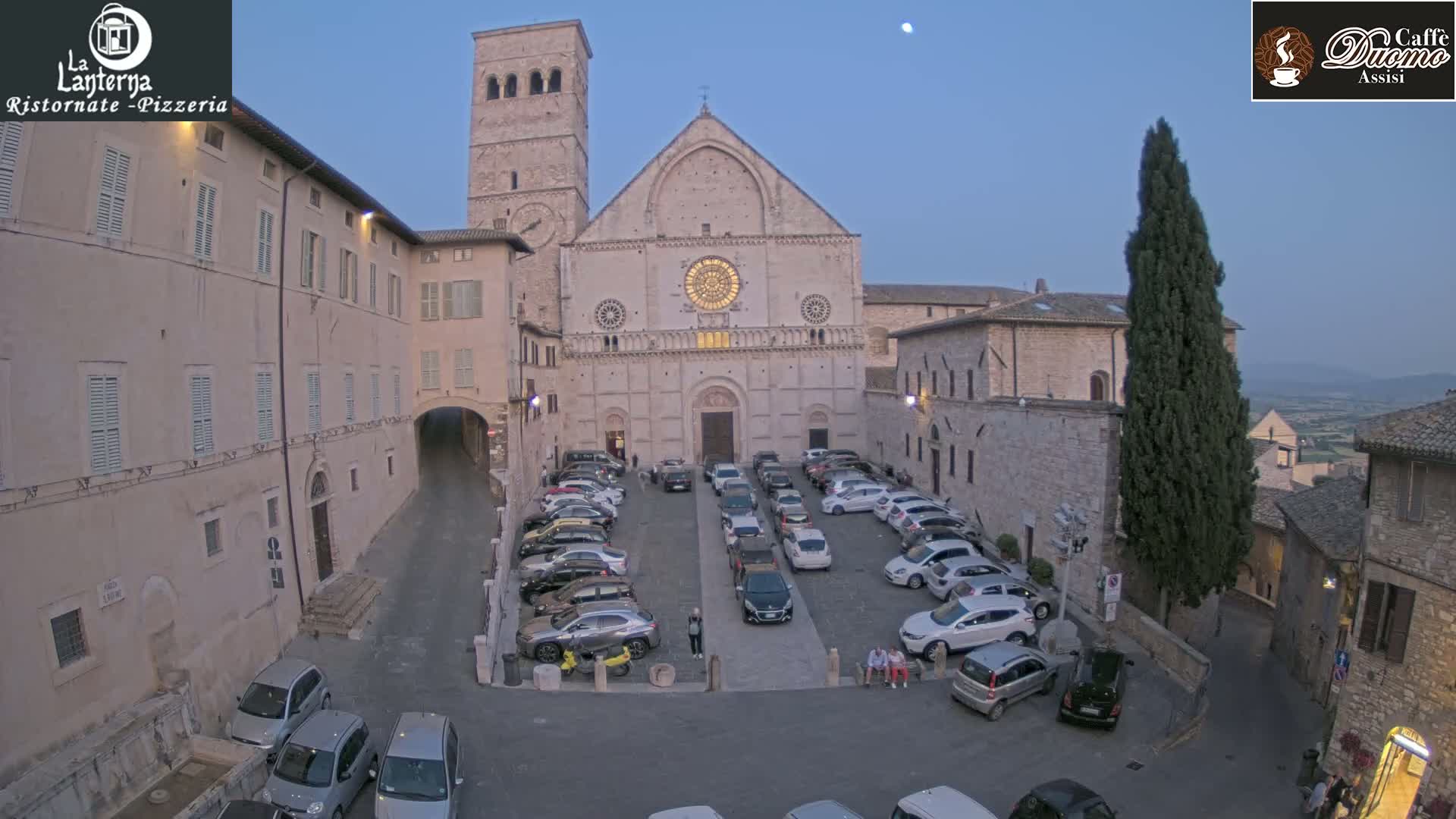A large stone church faces a town square filled with parked cars under a twilight sky.