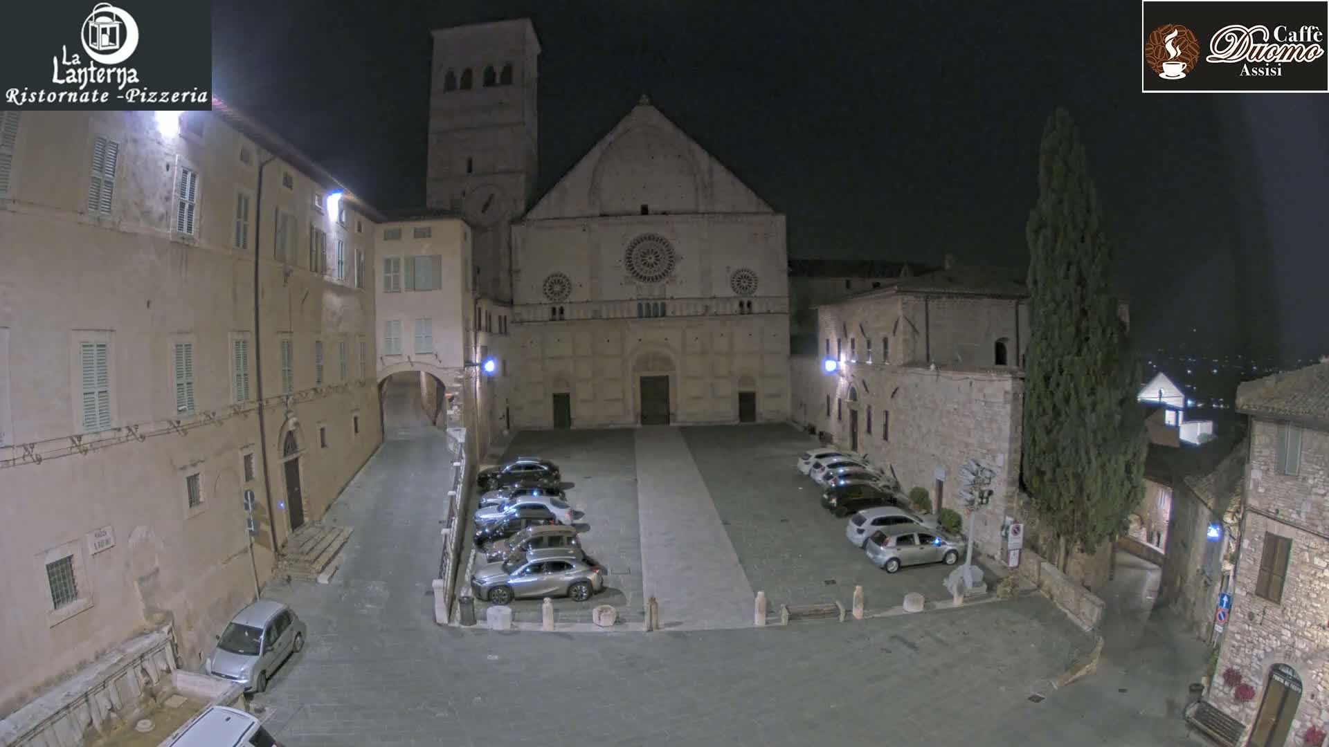 A nighttime view of a town square with cars parked in front of a large stone church, under a dark sky.