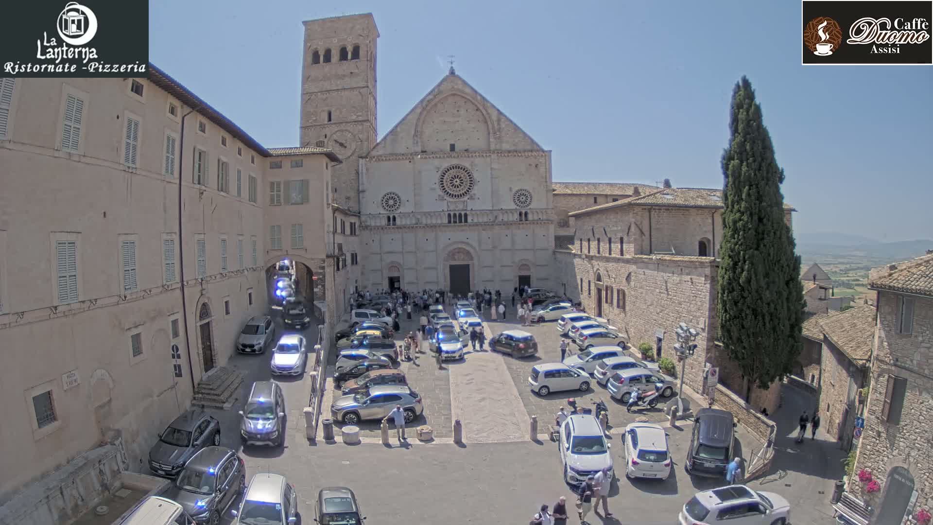 A large stone church is surrounded by a town square filled with parked cars and people on a sunny day.