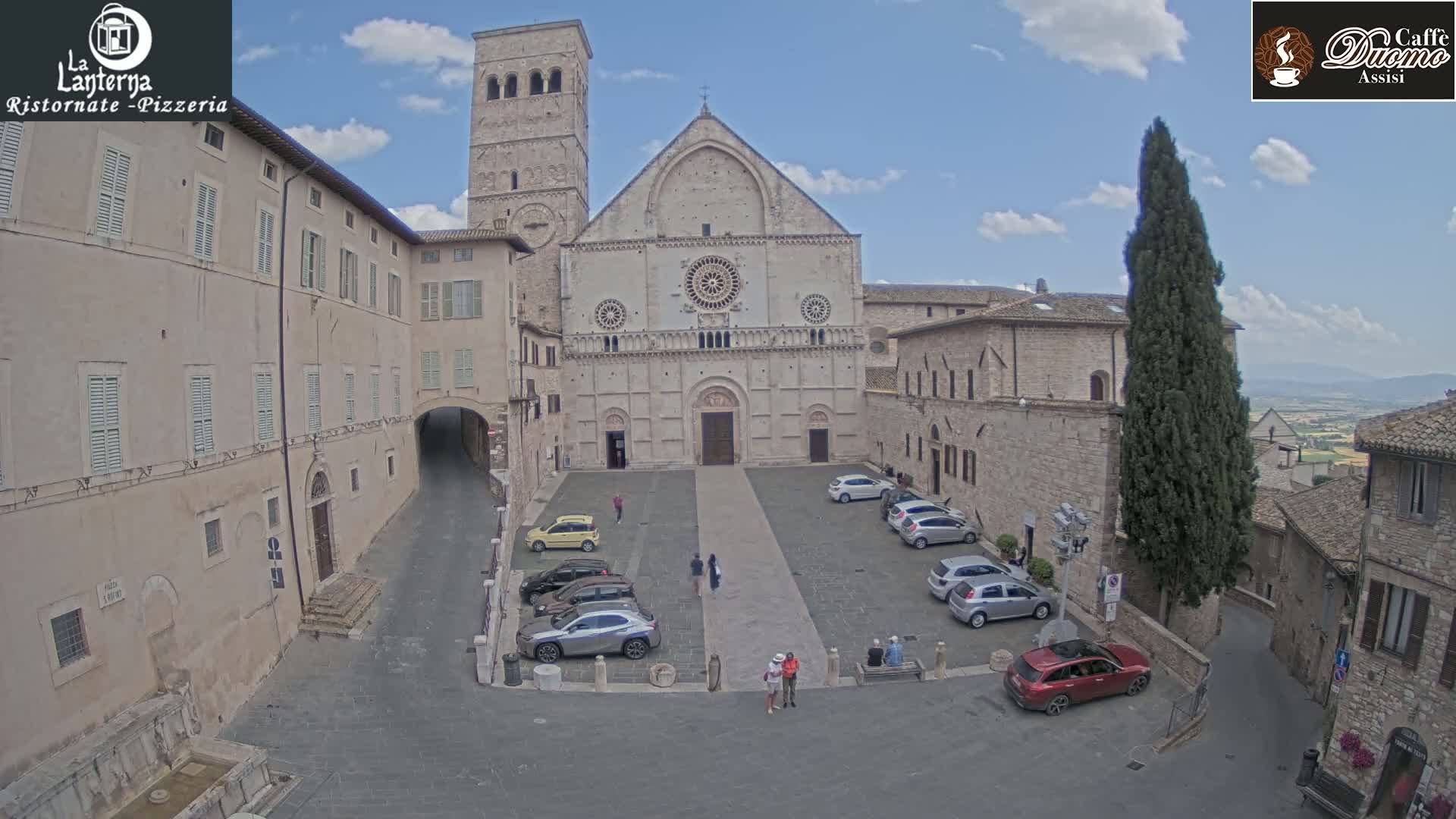 A large stone church stands in a town square with several cars parked, under a mostly sunny sky.