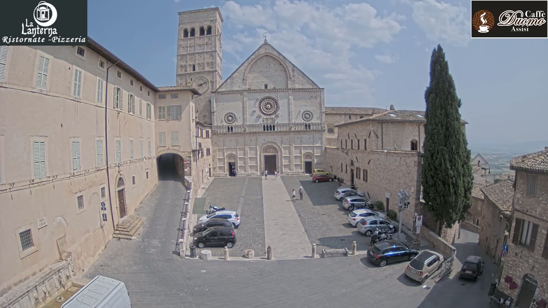 A large stone cathedral sits in a town square, surrounded by other stone buildings, cars, and a few pedestrians, under a mostly sunny sky.