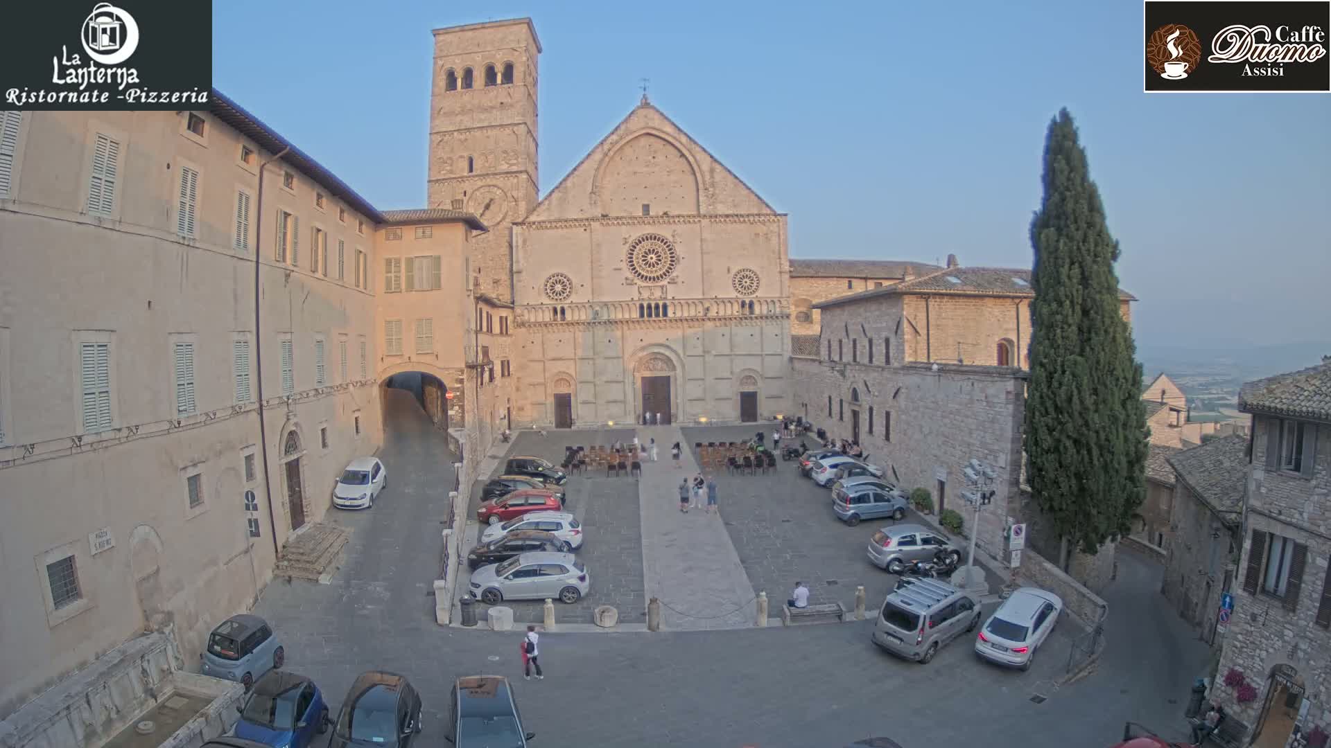 A large stone cathedral sits in a town square under a clear, light blue sky, surrounded by parked cars and a few pedestrians.