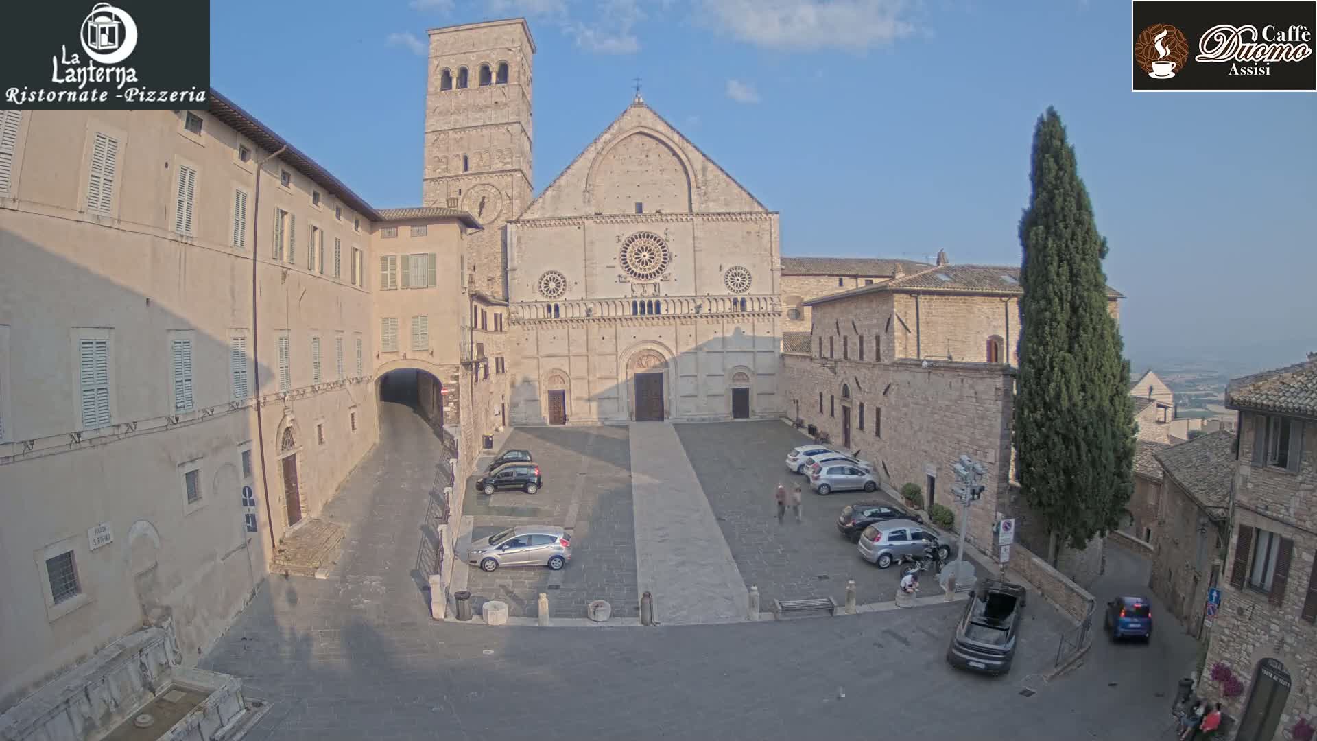 A large stone cathedral stands in a town square on a sunny day, surrounded by other stone buildings and a few parked cars.