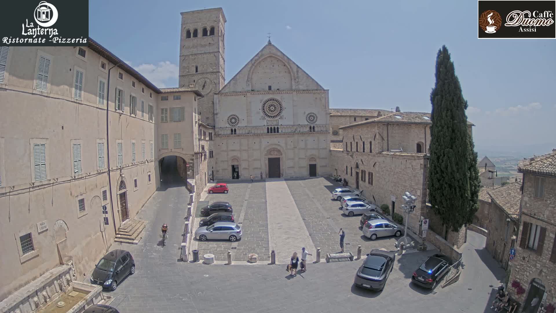 A sunny day reveals a town square with parked cars in front of a large stone church, surrounded by other stone buildings.