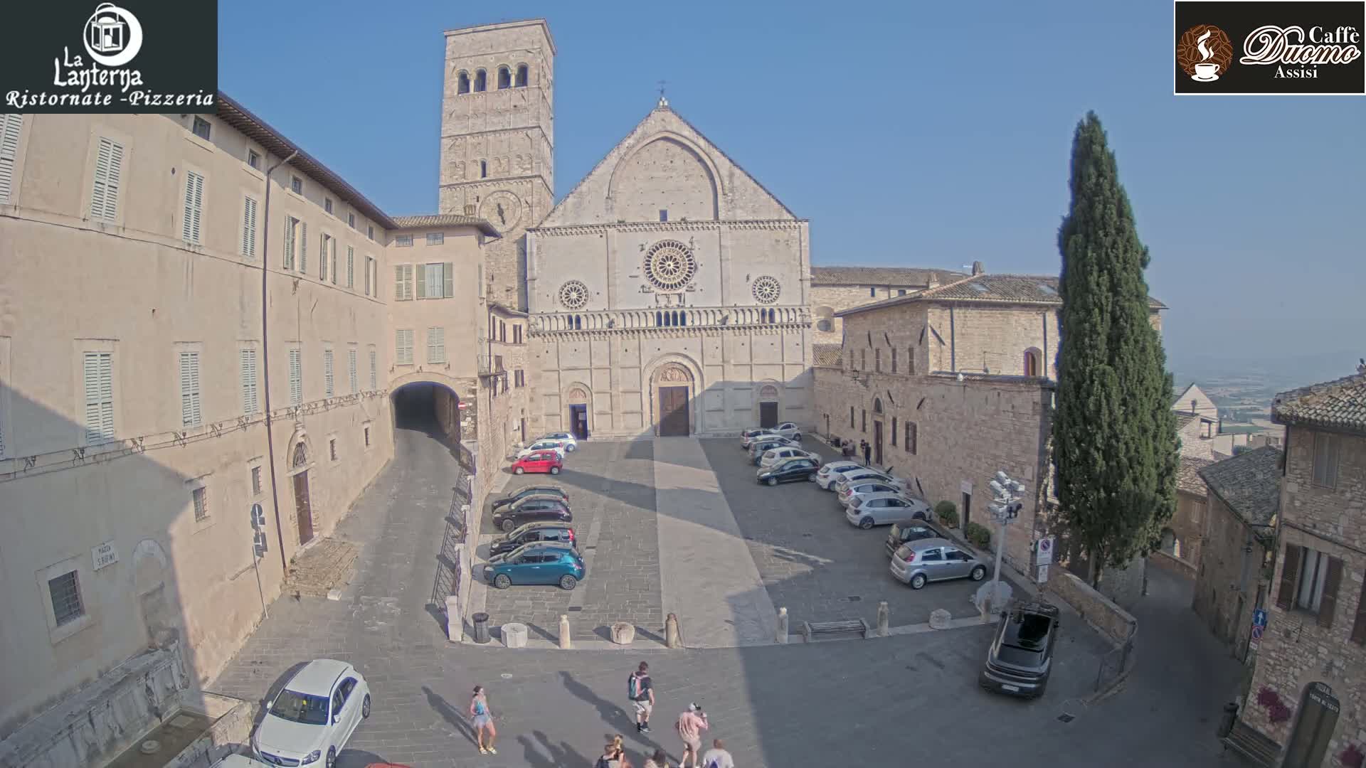 A large stone cathedral sits in a sunny town square, surrounded by buildings and a few parked cars, with several people walking nearby.