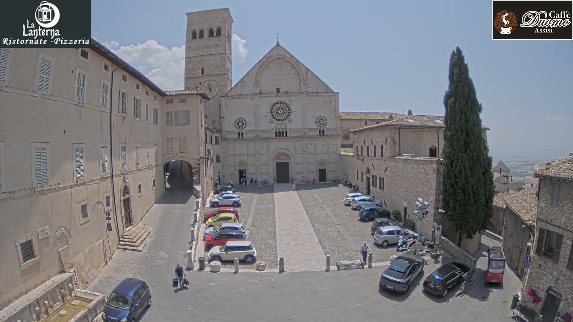 A large stone cathedral sits in a town square on a sunny day, with cars parked in the square and buildings surrounding it.