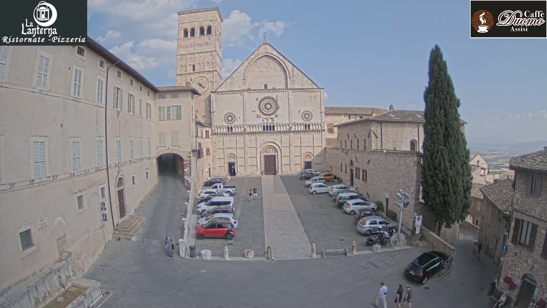A large, light-colored stone cathedral faces a square with parked cars under a mostly clear sky.