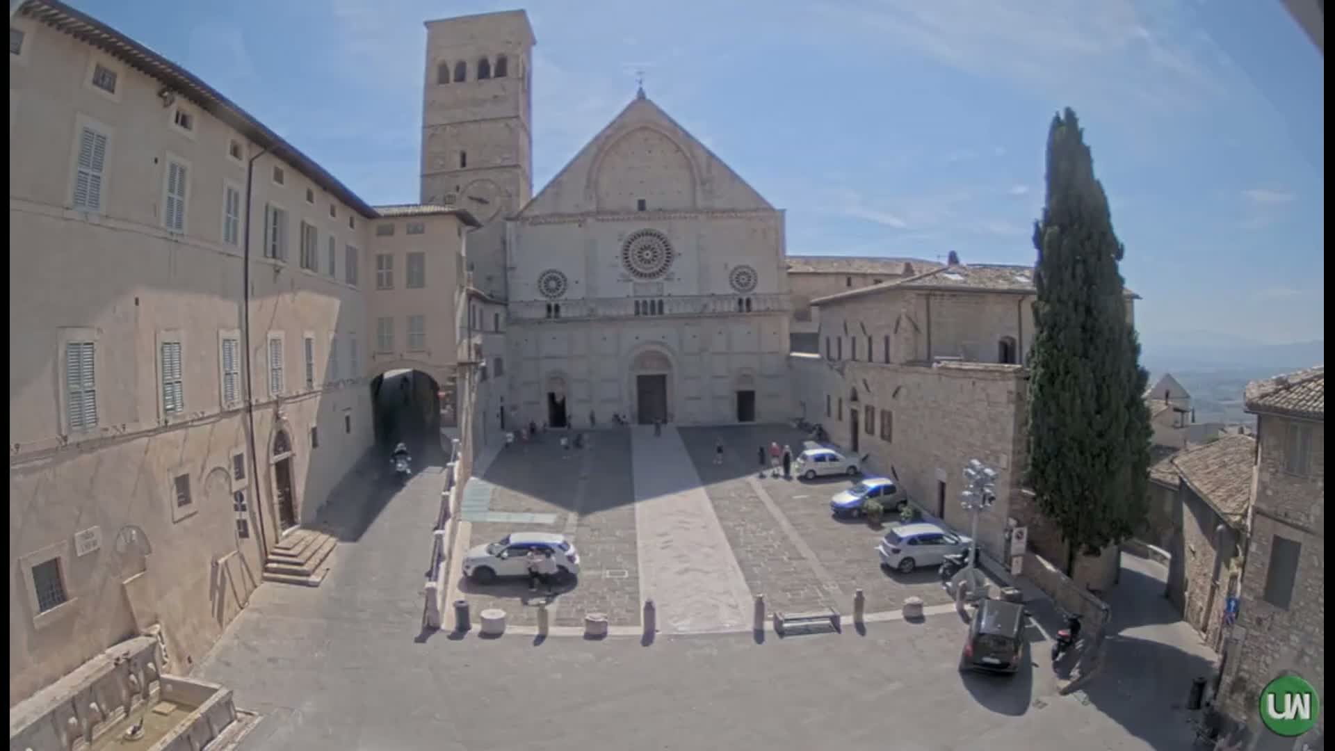 A large, light-colored stone cathedral stands in a sunny town square, surrounded by other stone buildings, with a few cars parked and some people strolling about.