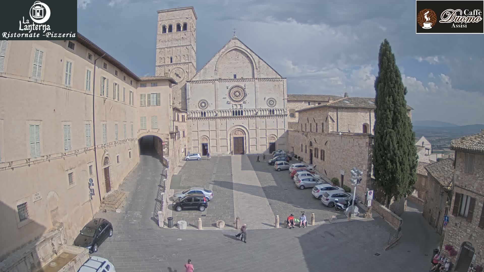 A partly cloudy day reveals a large stone church in a town square with several parked cars and a few pedestrians.