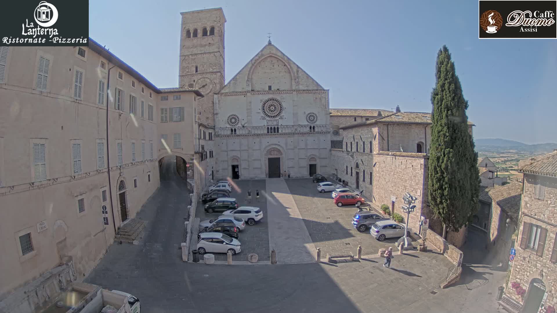 A large stone cathedral is situated in a town square with several cars parked around it, under sunny skies.