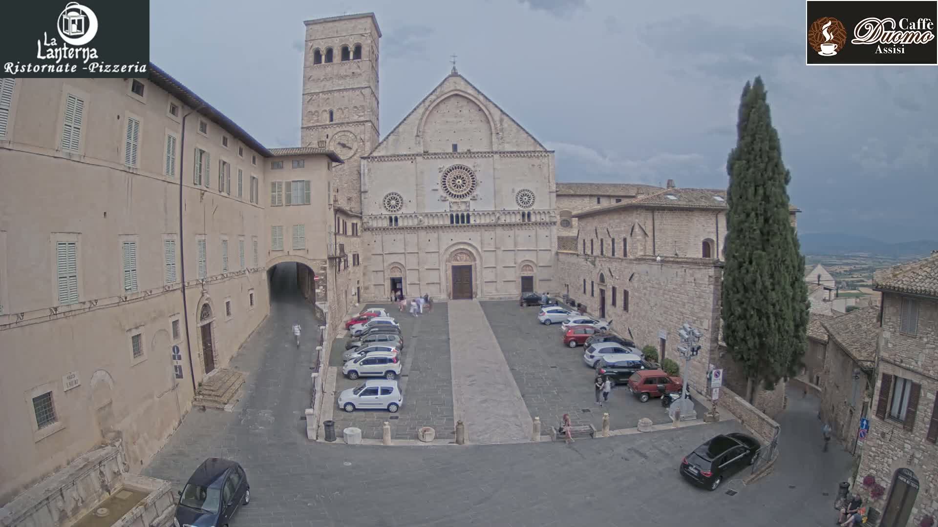 A large stone cathedral faces a town square with parked cars under an overcast sky.